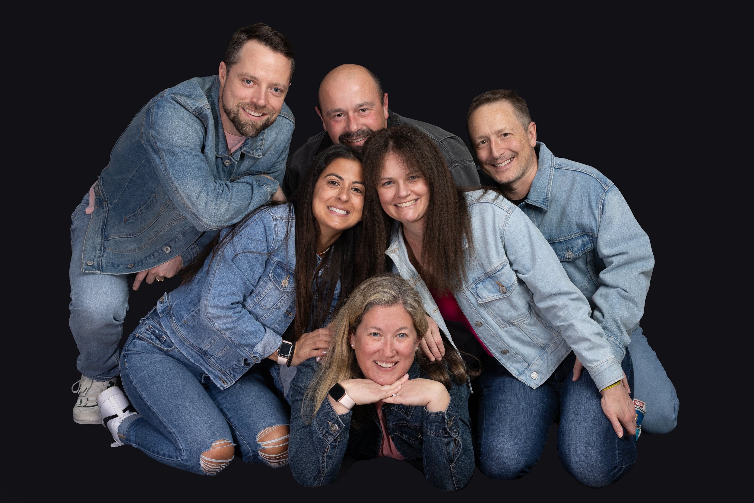 Group of six people posing in denim clothing on a dark background, all smiling.