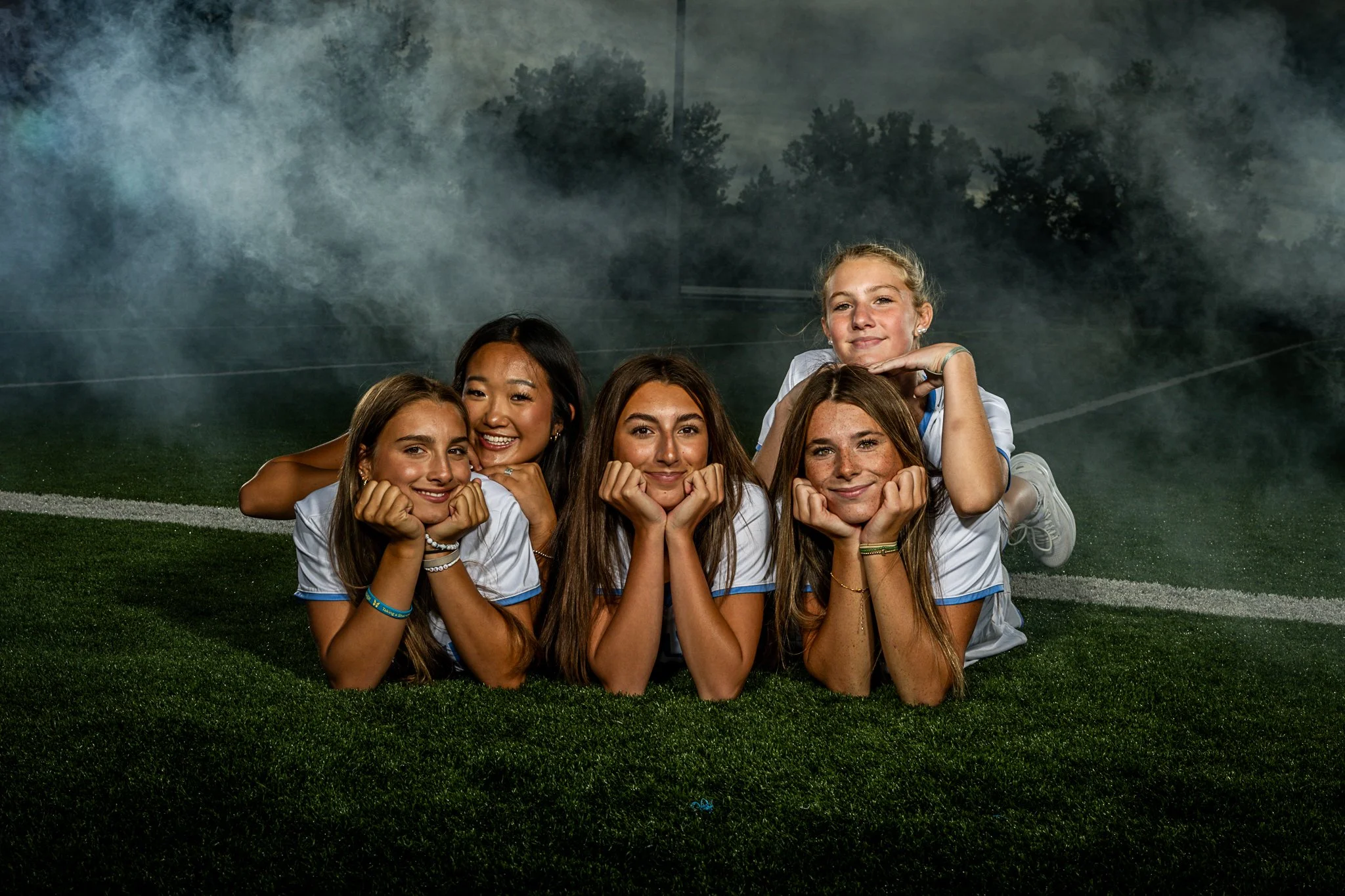 Group of five smiling young women lying on their stomachs on a grassy football field, propped up on elbows with chins resting on hands, with fog or smoke in the background