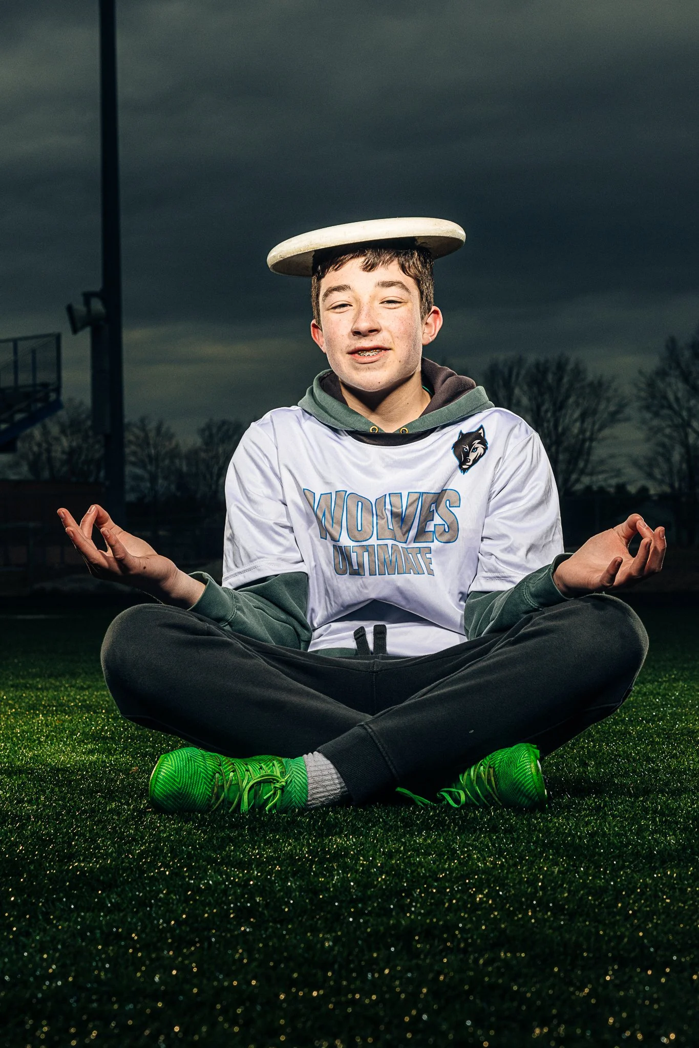 A young man sitting cross-legged on a sports field at dusk, balancing a frisbee on his head, wearing a white hoodie with 'WOLVES UNITE' lettering and green sneakers, with dark clouds in the background.
