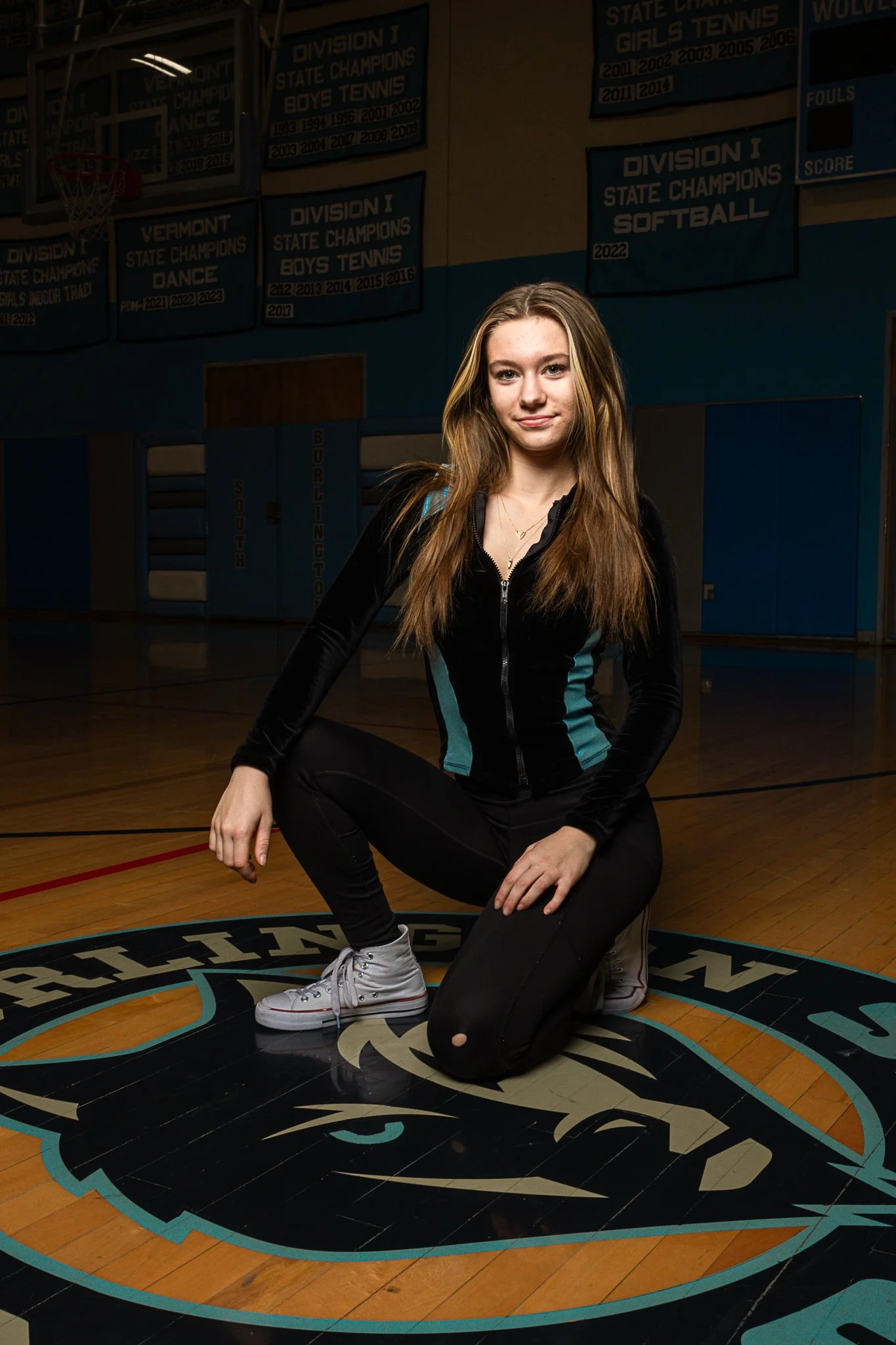 A young woman with long light brown hair, wearing a black zip-up jacket with turquoise accents, black pants, and white sneakers, kneeling on a gymnasium floor. She is on a logo with a lion's head inside a circle, with banners hanging in the backgroun