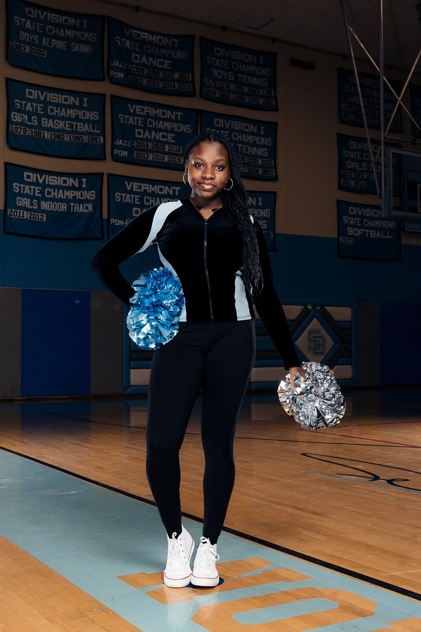 Young woman standing on a gymnasium basketball court holding blue and silver cheerleading pom-poms, background features banners celebrating Vermont state championships.