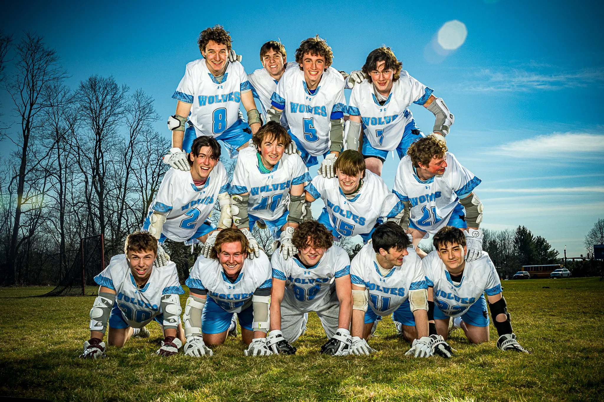 A group of young male hockey players in white and blue uniforms, posing in a pyramid formation on a grassy field, with leafless trees and a blue sky in the background.