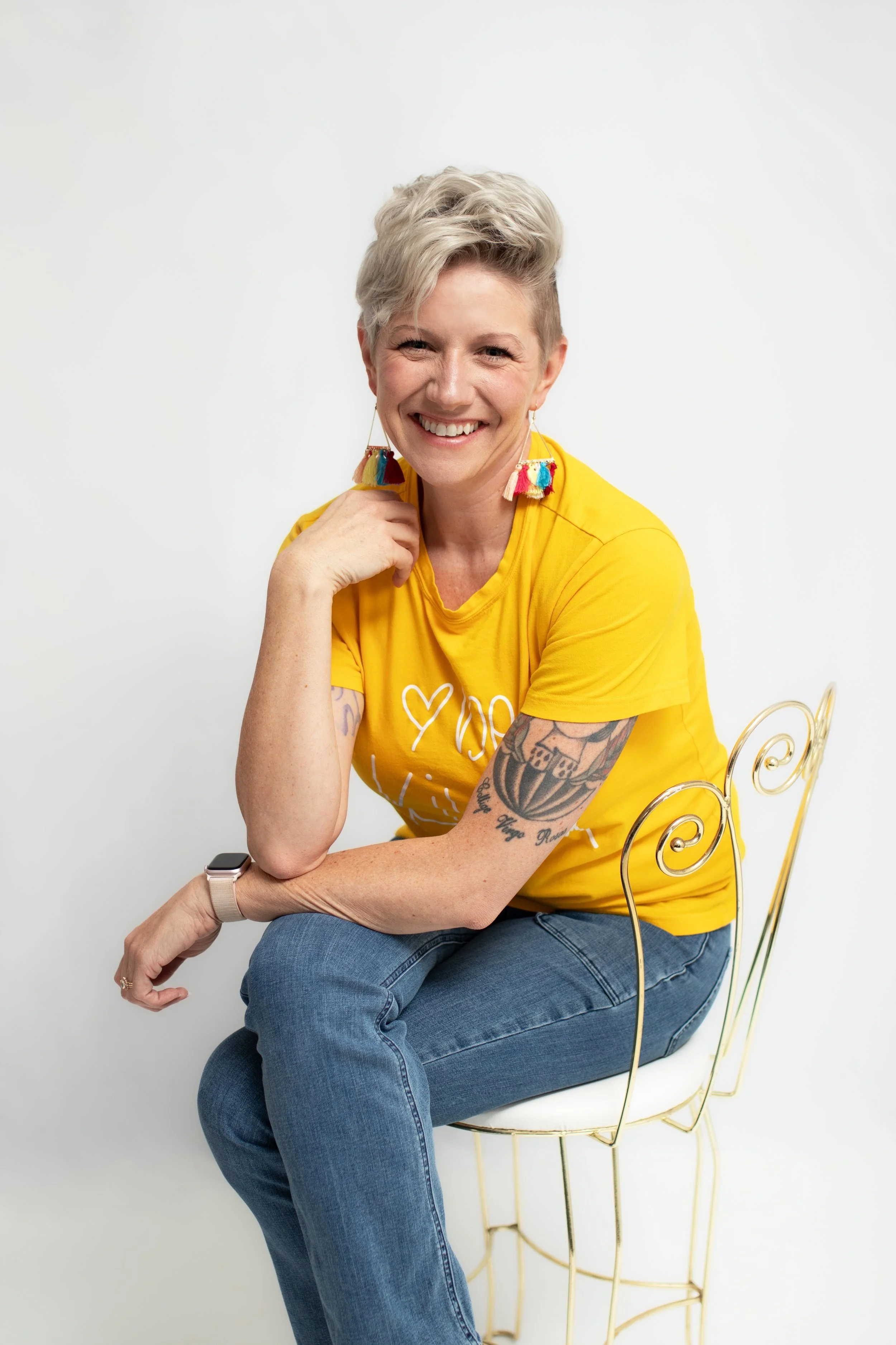 Business Coach Michelle Smock smiles warmly at the camera wearing a bright yellow tee shirt that says "be kind"