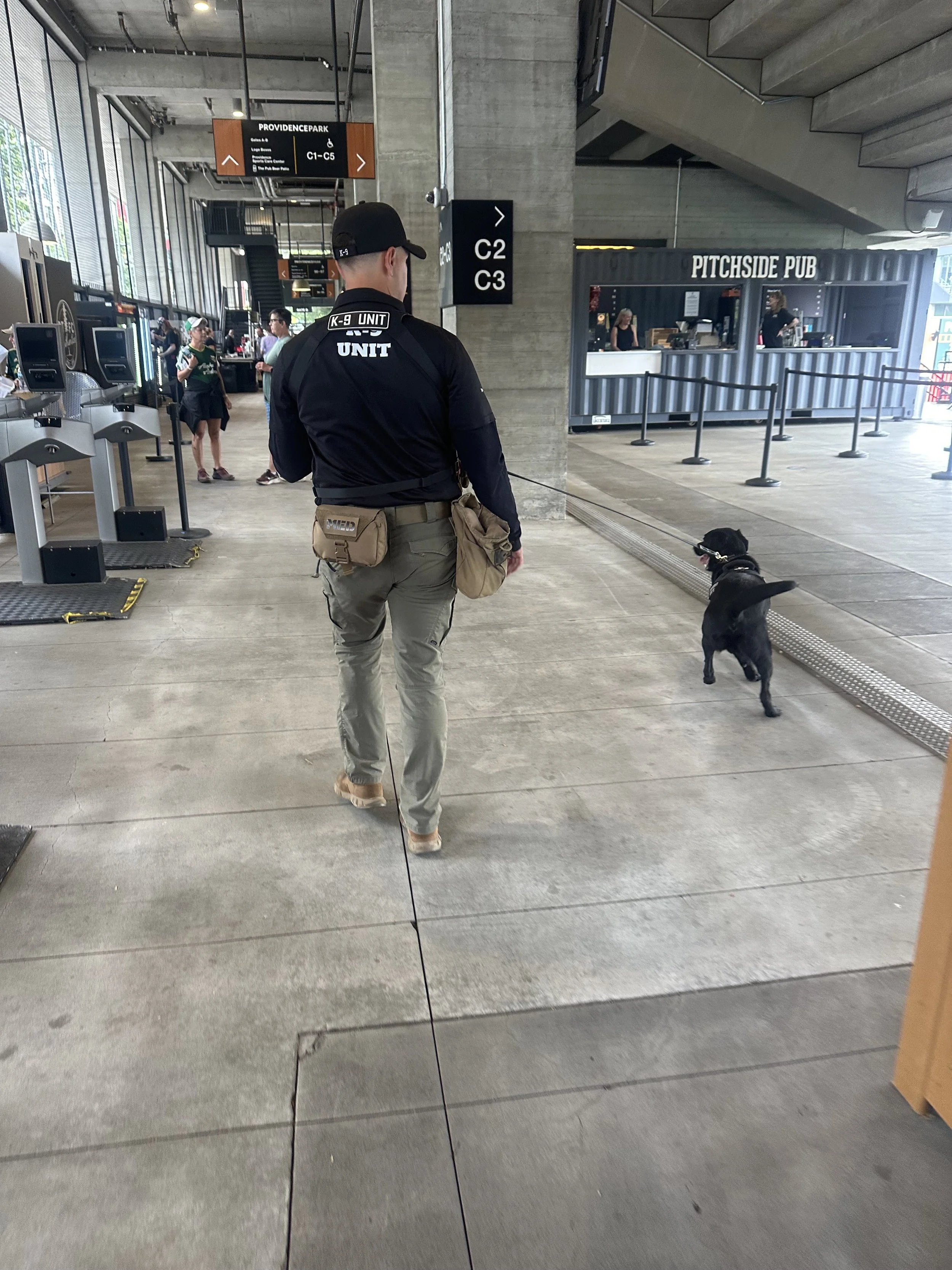A man walking a black service dog inside a modern airport terminal near a sign that reads Providence Park, with a pub in the background.