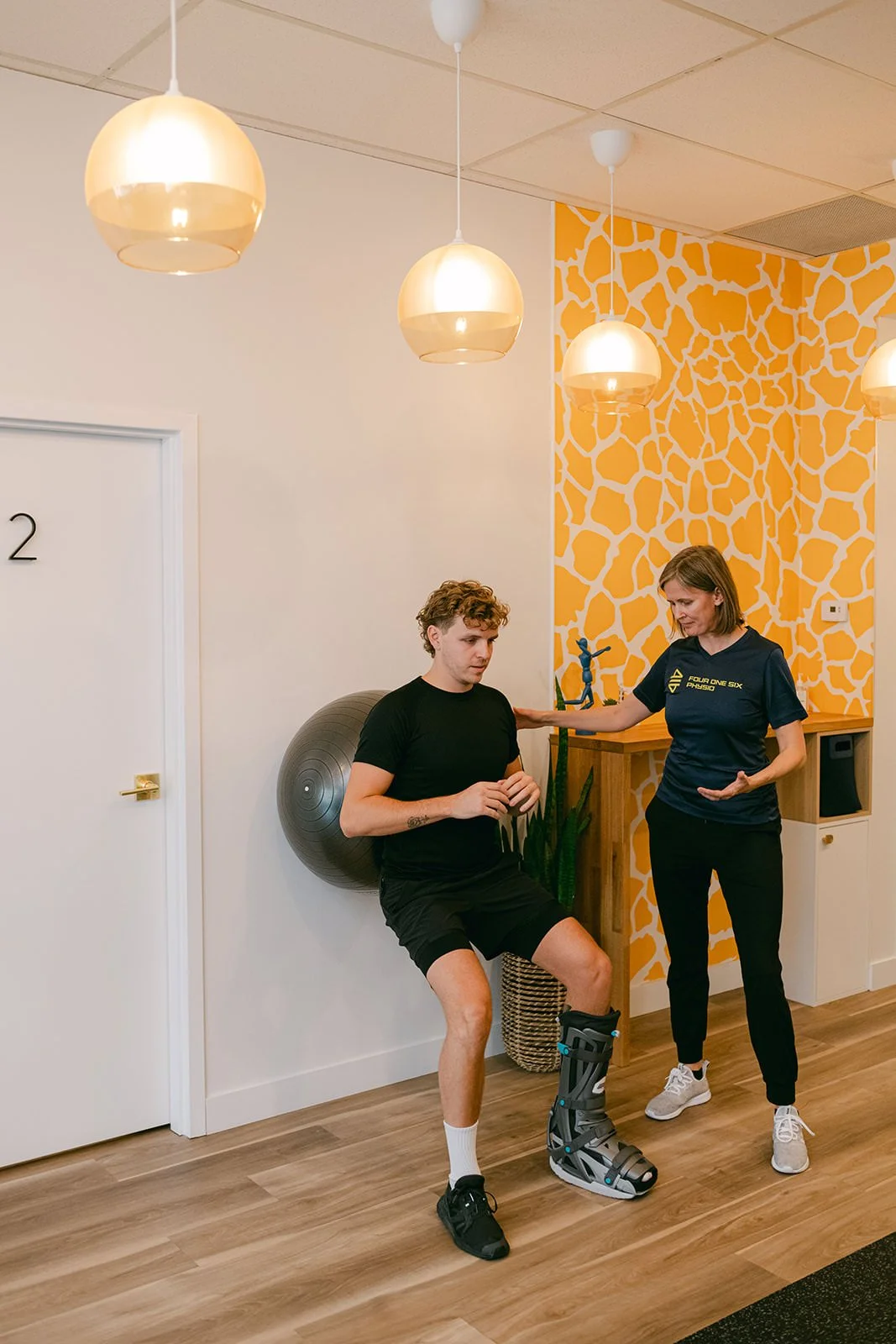 Physiotherapist supervising a patient performing an early rehabilitation exercise while wearing a walking boot after an Achilles tendon rupture.