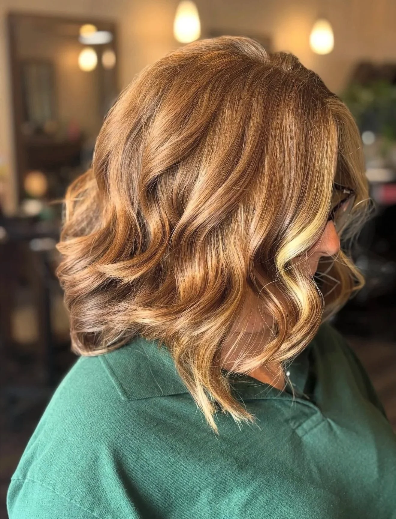 Woman with shoulder-length, wavy, blonde hair wearing glasses and a green top in a salon.