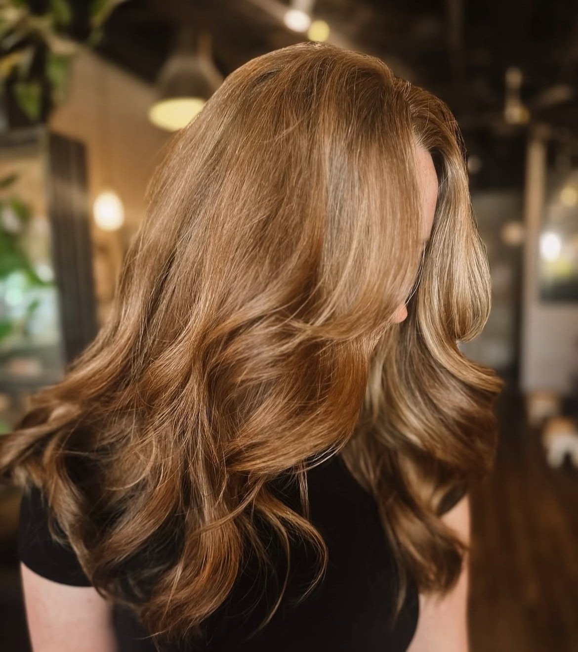 Close-up of a woman with long, wavy, light brown hair, looking downward in a dimly lit indoor setting.
