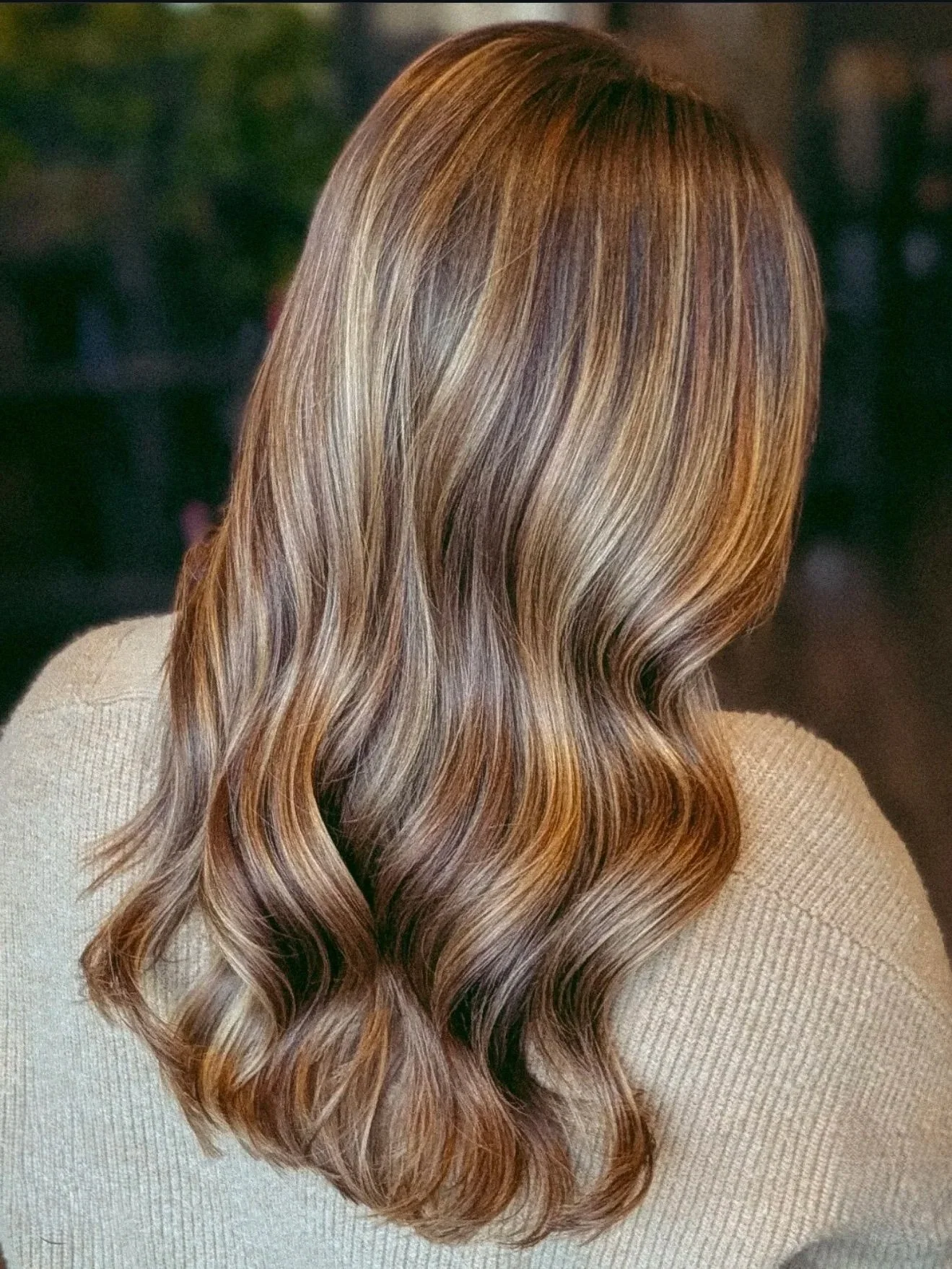 Back view of a woman with long wavy hair in shades of blonde and light brown, wearing a beige sweater.