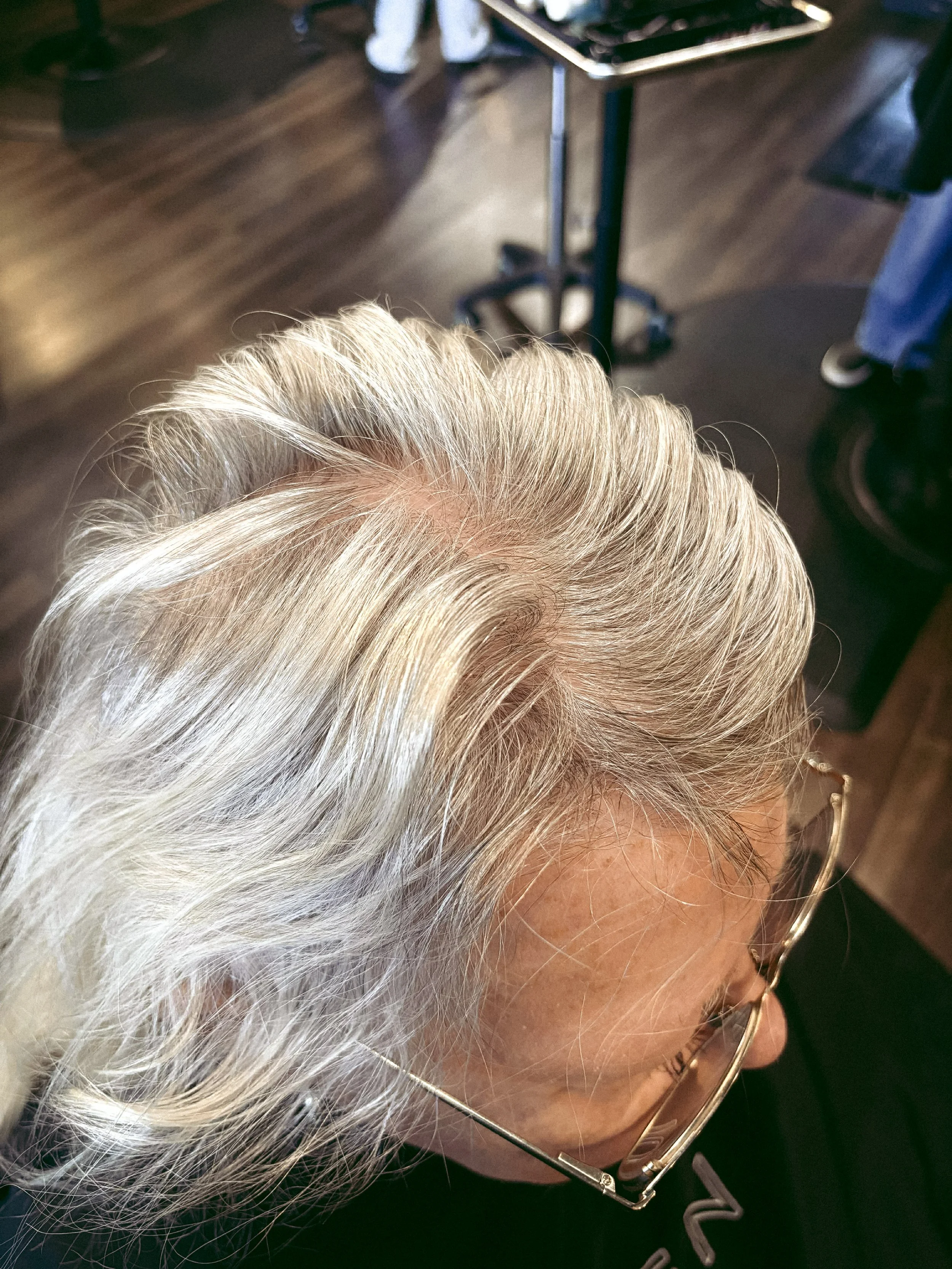 Close-up of a senior woman with white hair, wearing glasses, in a salon with wooden flooring and a hairdresser's station in the background.
