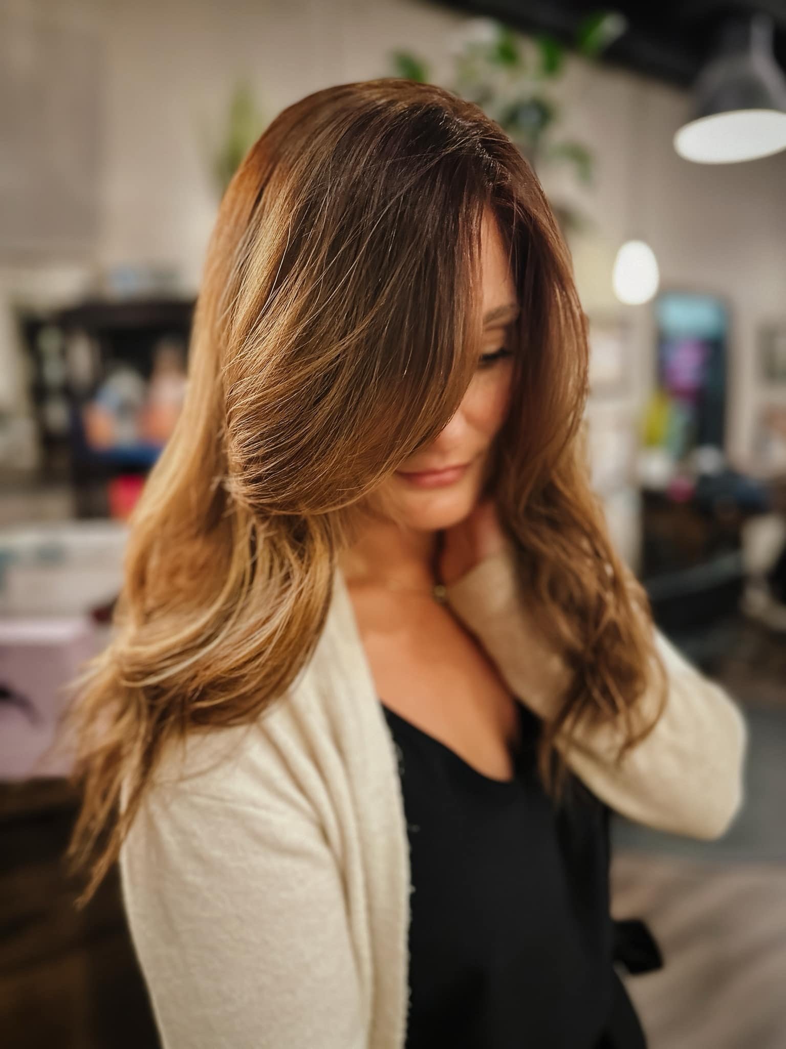 A woman with long, wavy, light brown hair looking down, wearing a beige cardigan and black top, in an indoor setting with blurred background.