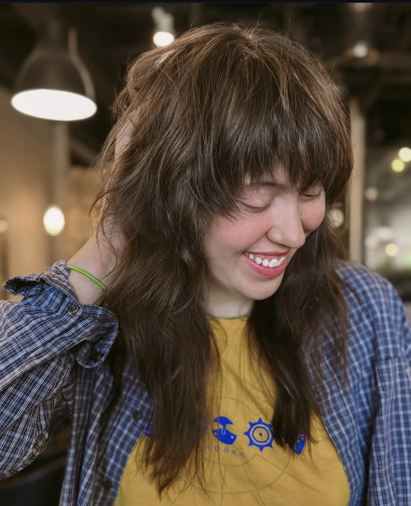 A woman smiling with her eyes closed, wearing a yellow shirt and a blue plaid shirt, in a warmly lit indoor setting.