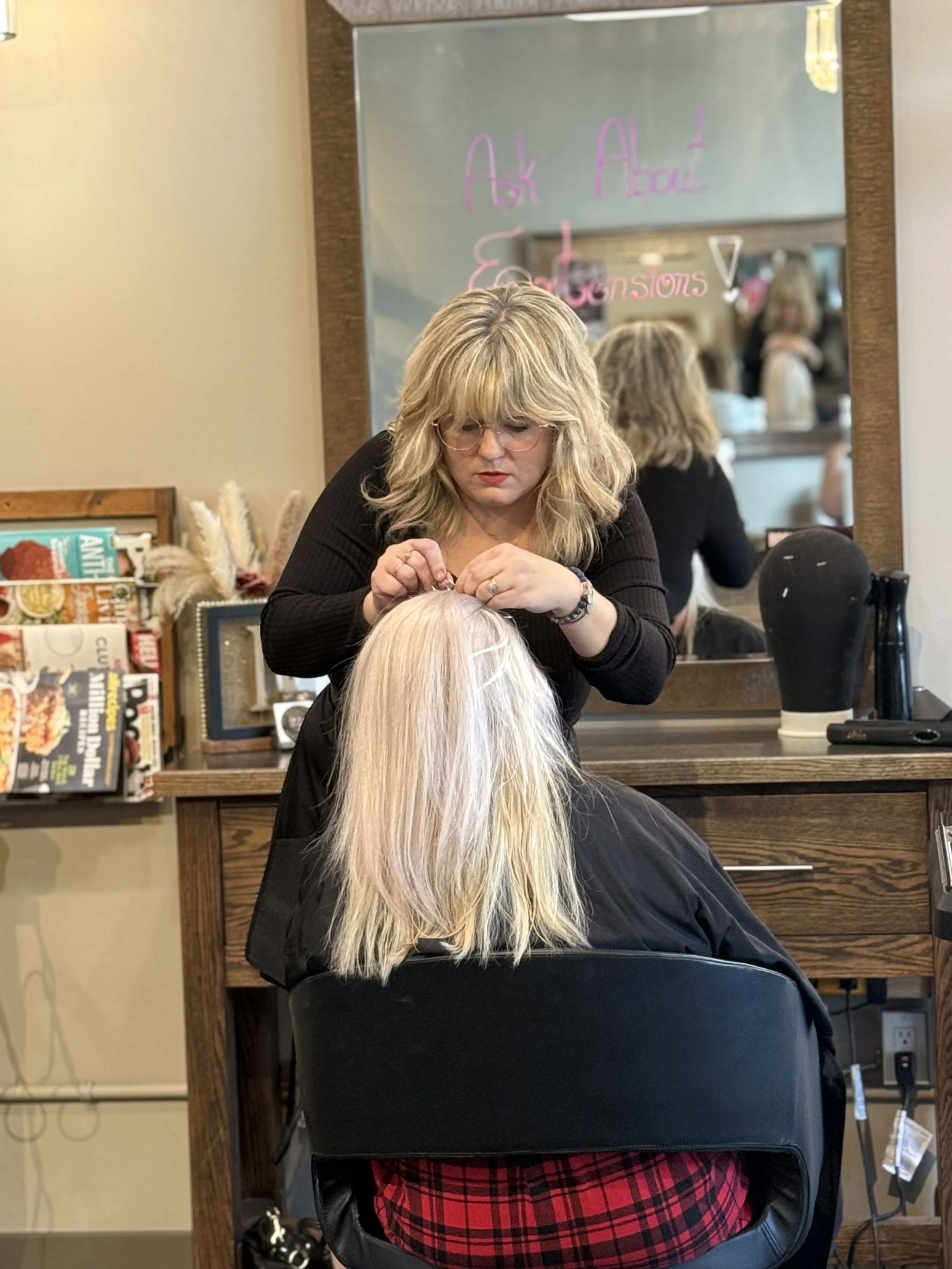 A woman with blonde hair, wearing glasses and a black top, is styling a client's long, blonde hair at a salon. The client is sitting in a black chair with a red plaid skirt visible. There is a mirror behind them reflecting the scene and background elements include a wooden desk with magazines and decorations, and a large mirror with pink writing that says 'Ask About Extensions!'