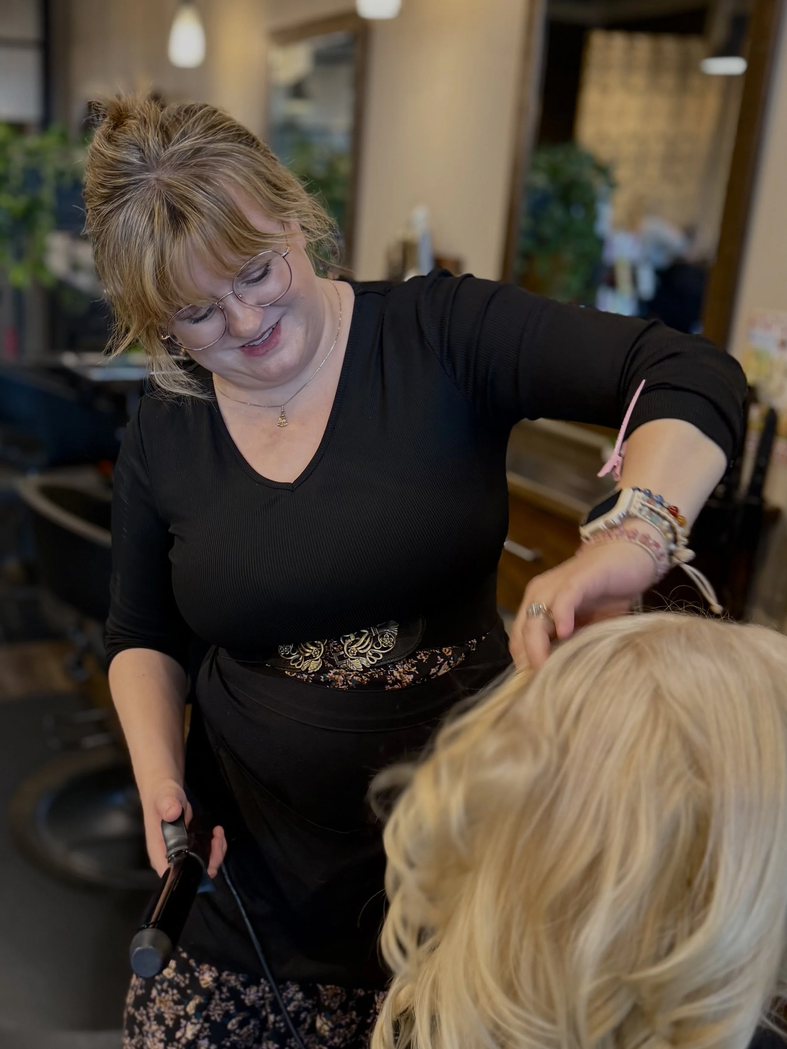 A hairstylist with glasses and a black top is smiling while working on a client's blond hair in a salon.