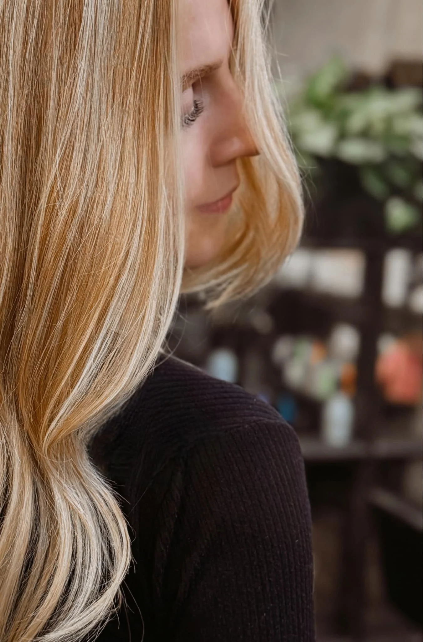 A woman with long blonde hair in profile, wearing a dark top, in an indoor setting with shelves and blurred objects in the background.