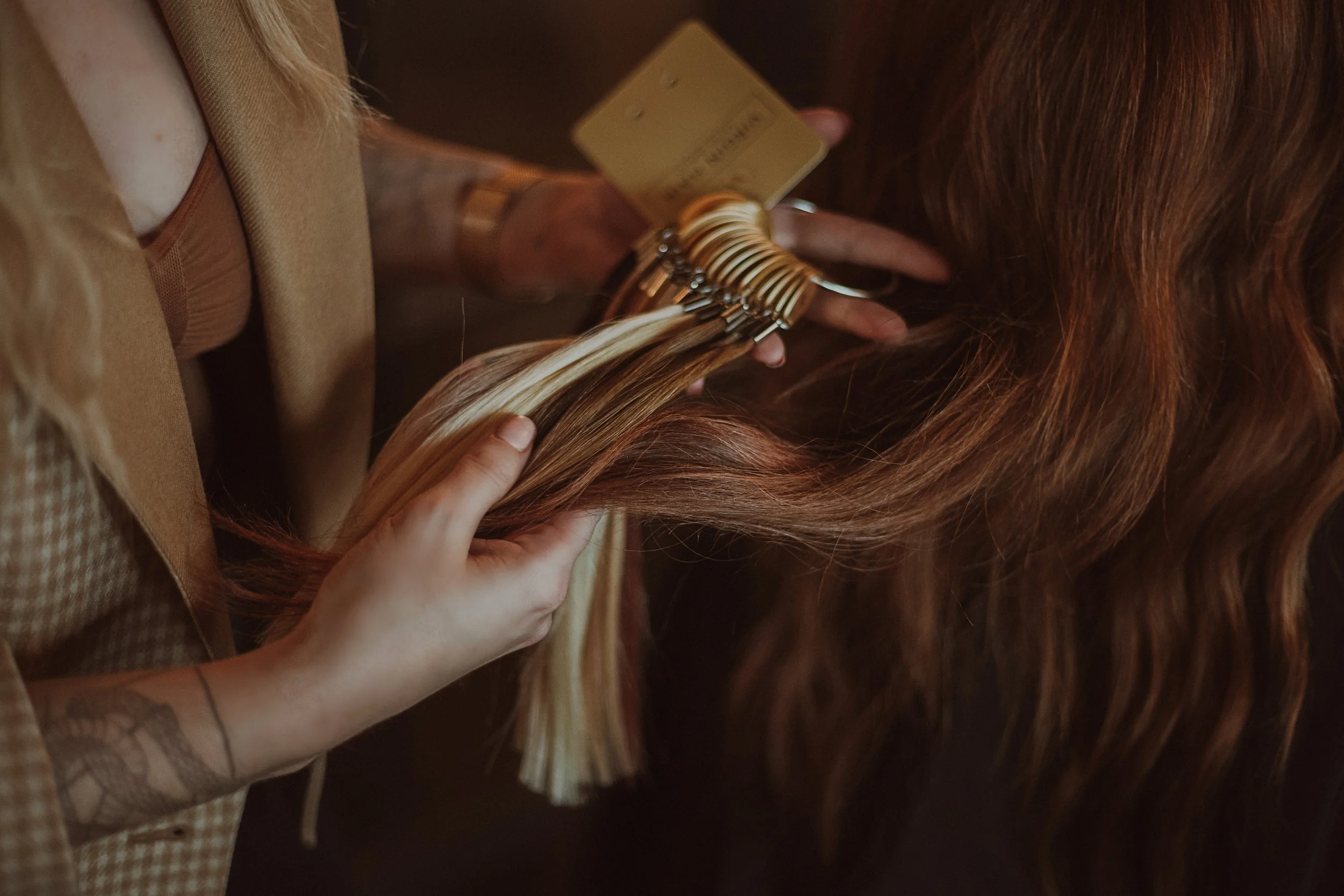 A hairdresser holding blonde hair extensions with a finger, displaying a hair color sample card, and a person with long auburn hair is sitting nearby.