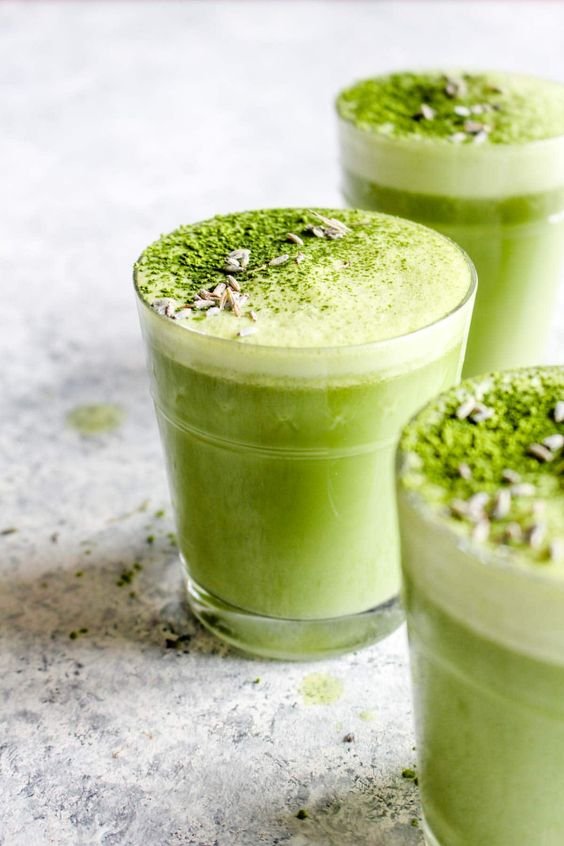 Glasses of green matcha latte topped with matcha powder and seeds on a light surface.