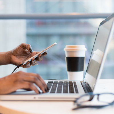 Person using a smartphone and working on a laptop at a desk, with a to-go coffee cup nearby.