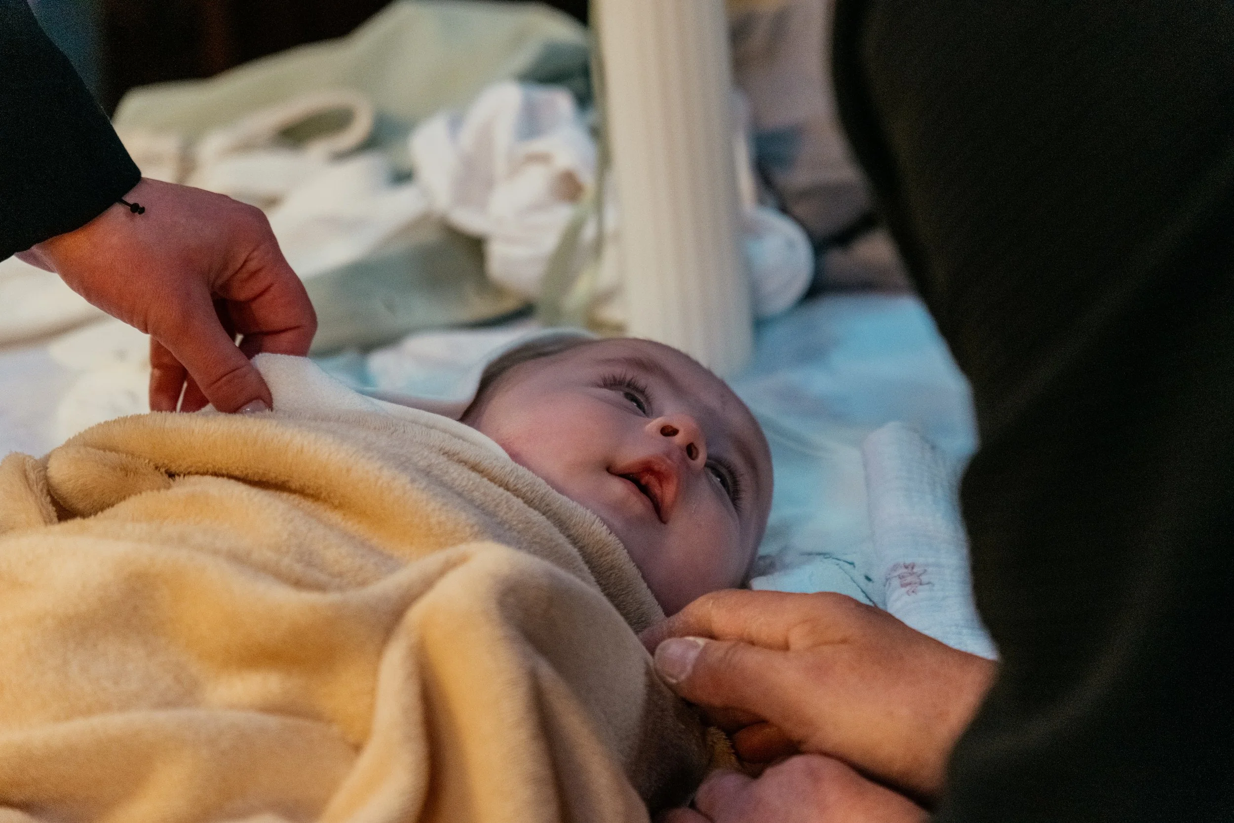 A baby lying on a bed, looking up at someone holding their hand, with soft blankets and bedding in the background.