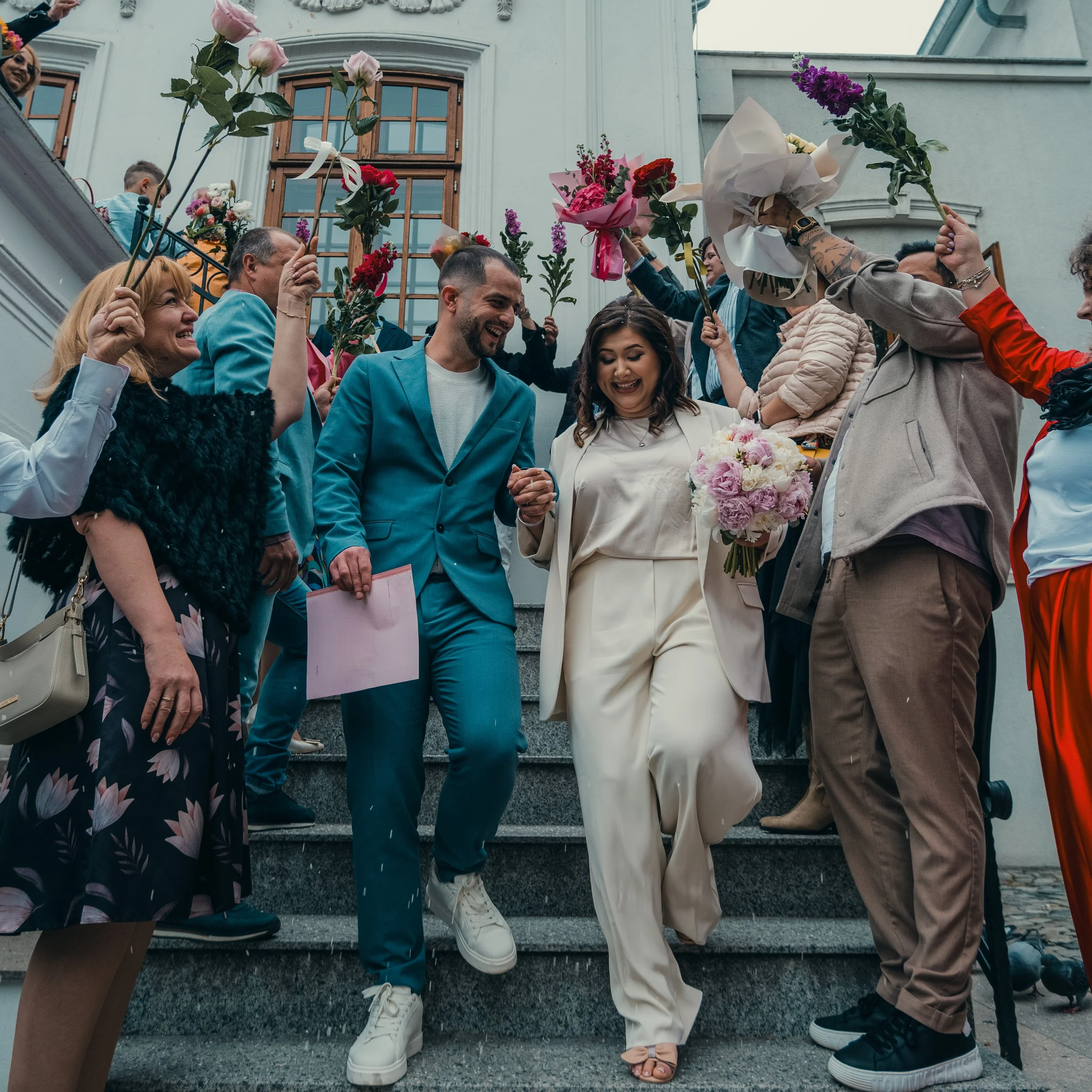 People holding bouquets of flowers while standing on outdoor steps near a building with large windows.