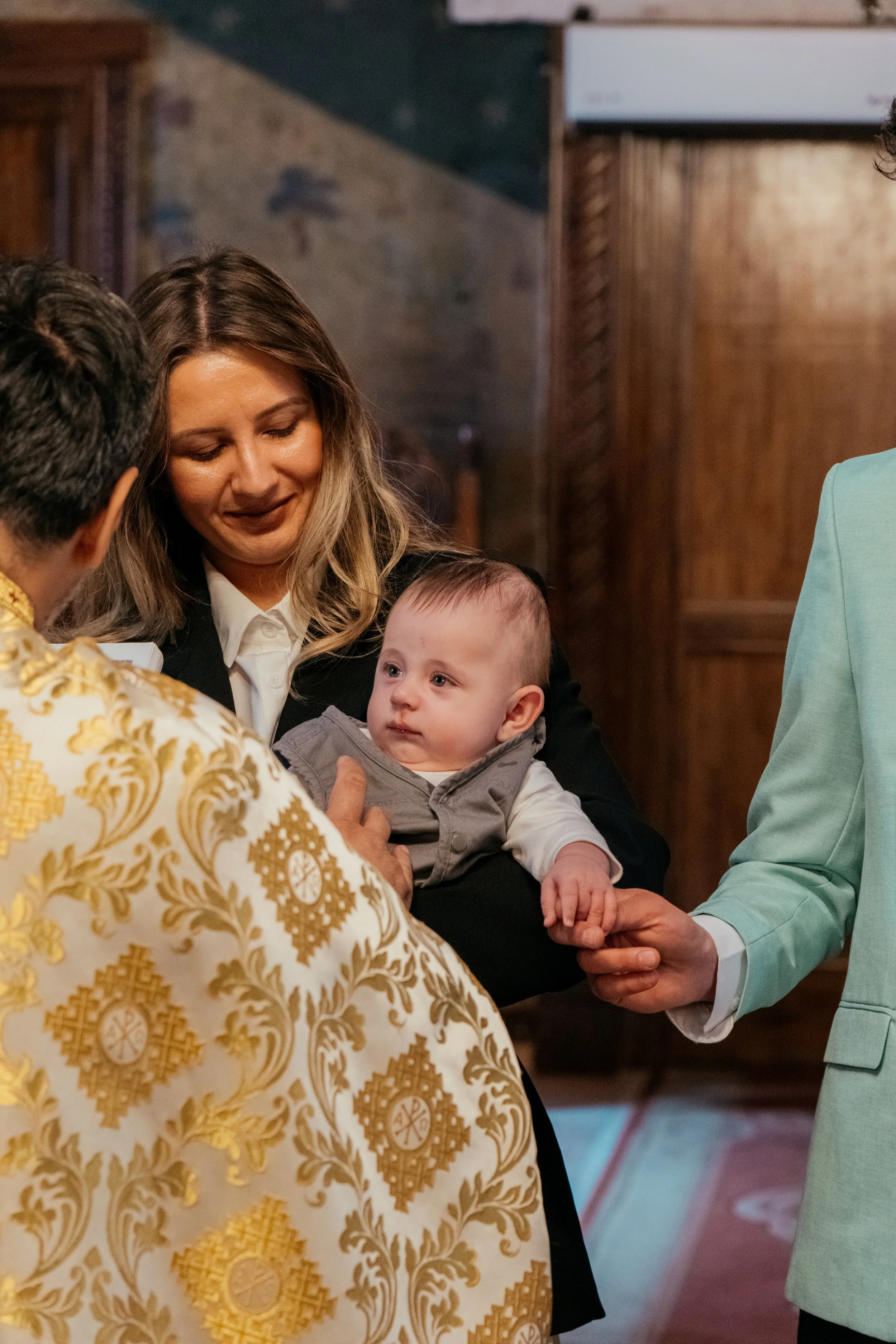 A woman holding a young child in her arms while standing next to a man. The woman has long, wavy hair and is dressed in dark clothing, while the child is wearing a gray jacket. The man has short dark hair and is wearing a colorful shirt with gold pat