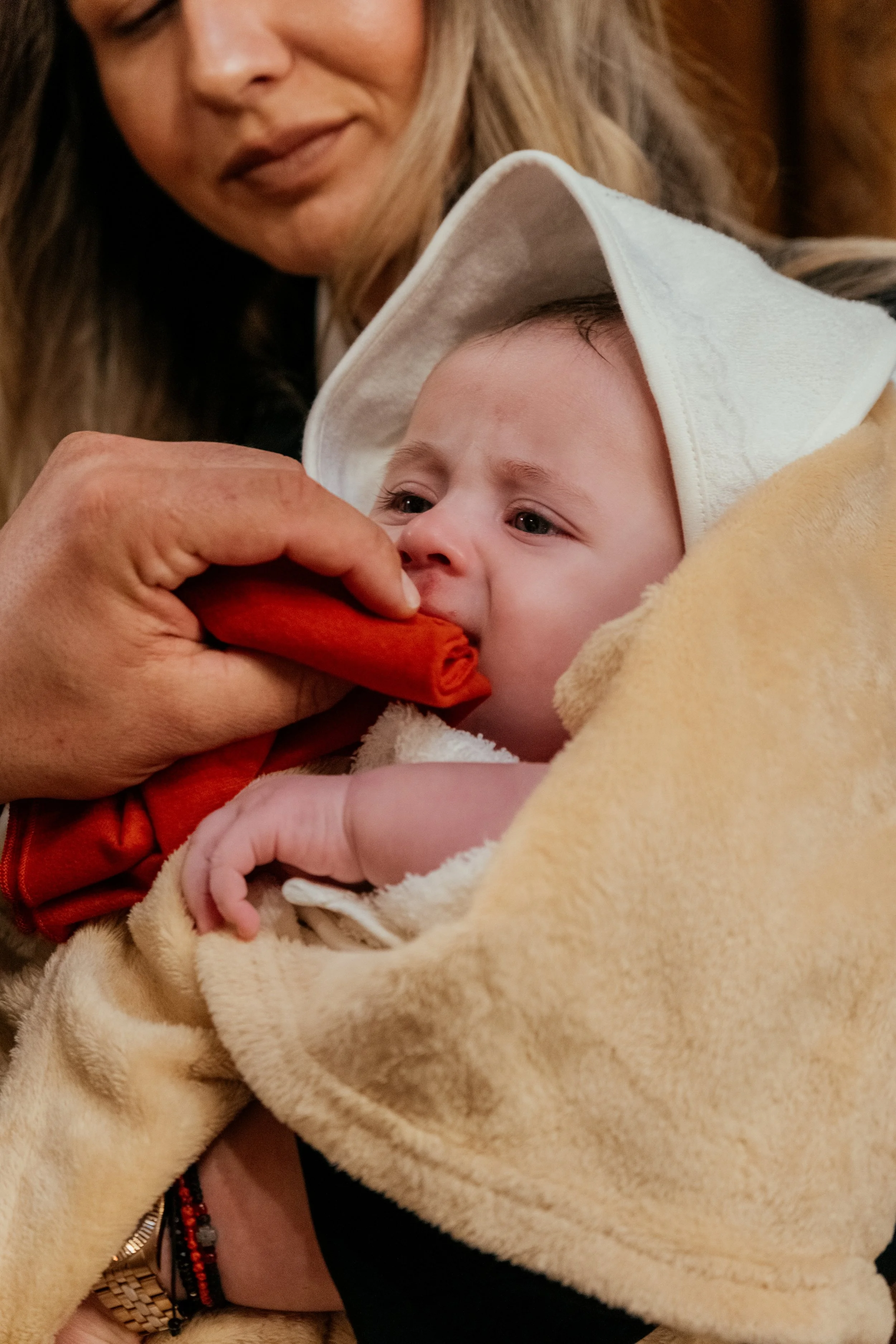 A woman with long blonde hair holding a baby wrapped in a beige blanket. The baby is wearing a white hooded towel or hoodie and is feeding on a rolled-up cloth or blanket. The woman is smiling gently, and the baby appears to be looking at the cloth.