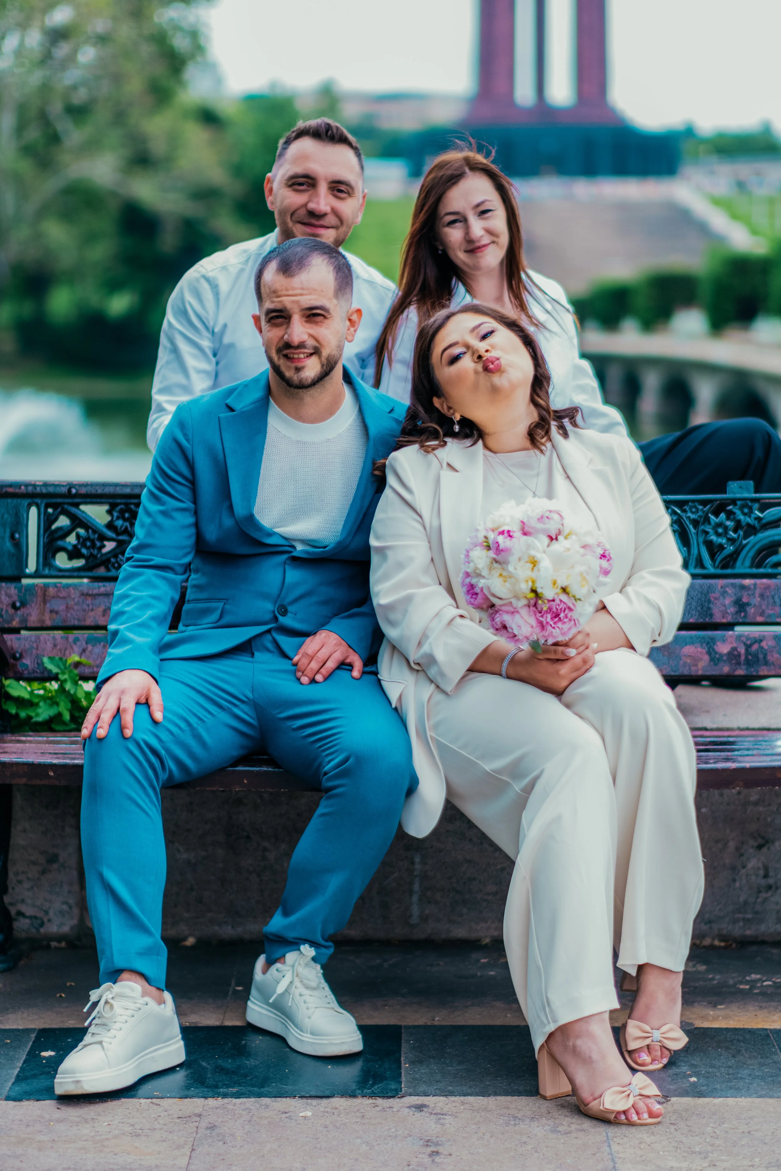 Four friends sitting on a park bench, with a bridge and greenery in the background. The group includes two men and two women, one woman is holding a bouquet of flowers, and they are all smiling and posing for the photo.