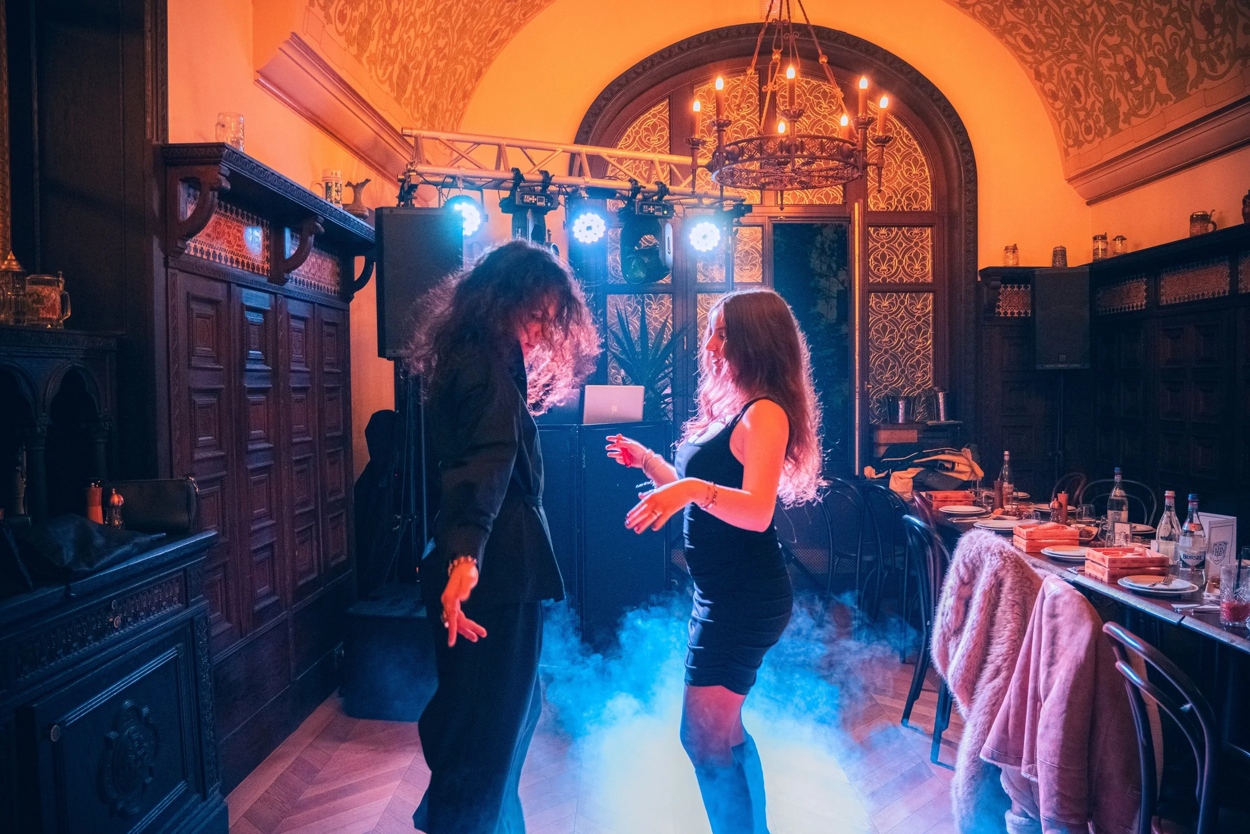 Two women dancing and talking inside a dimly lit, ornate room with wooden paneling, a chandelier, and a table with glasses and food.