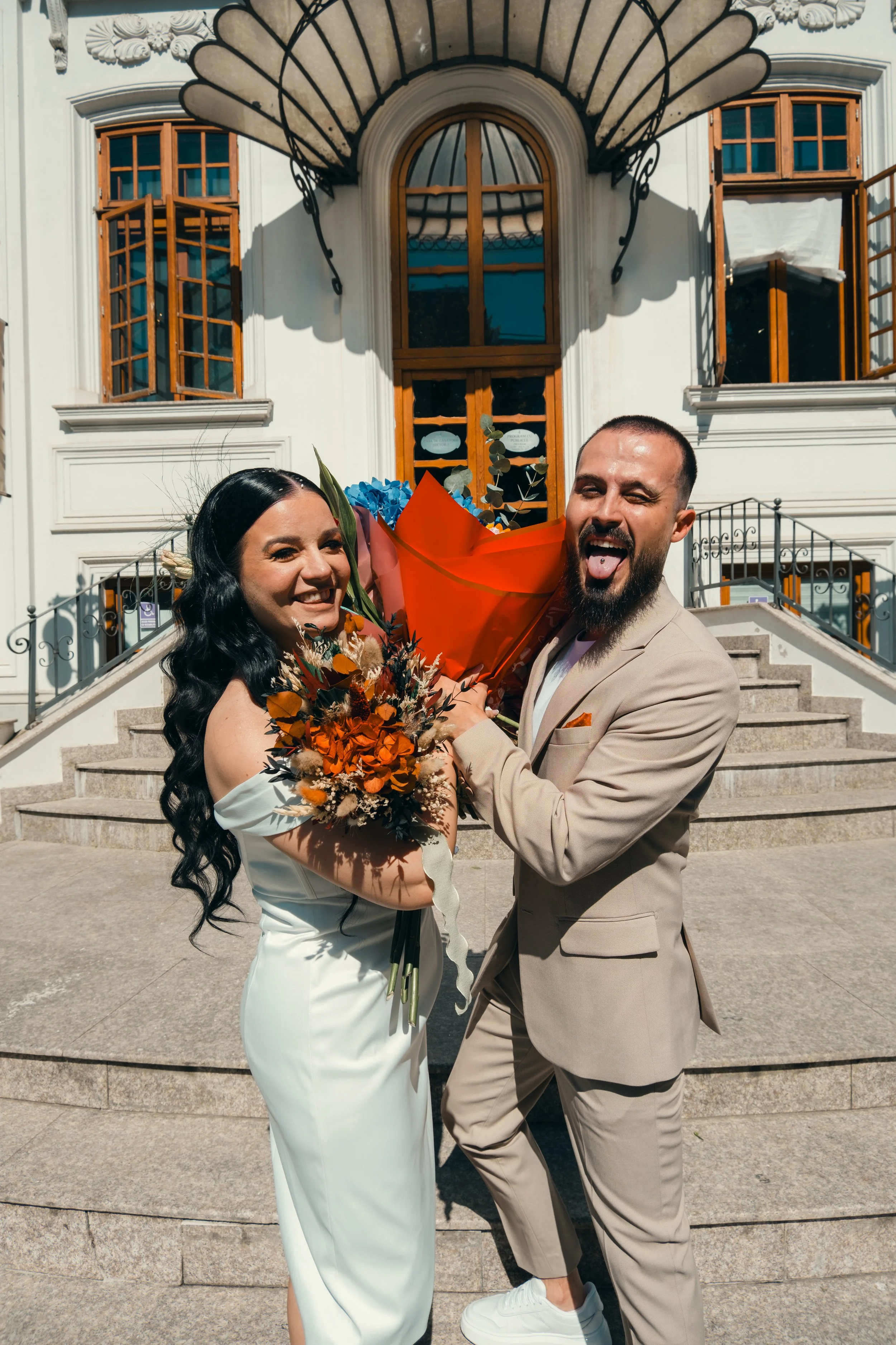 A joyful couple standing on the steps outside a grand white building with large wooden framed windows, holding a bouquet of flowers, smiling and sticking out their tongues.