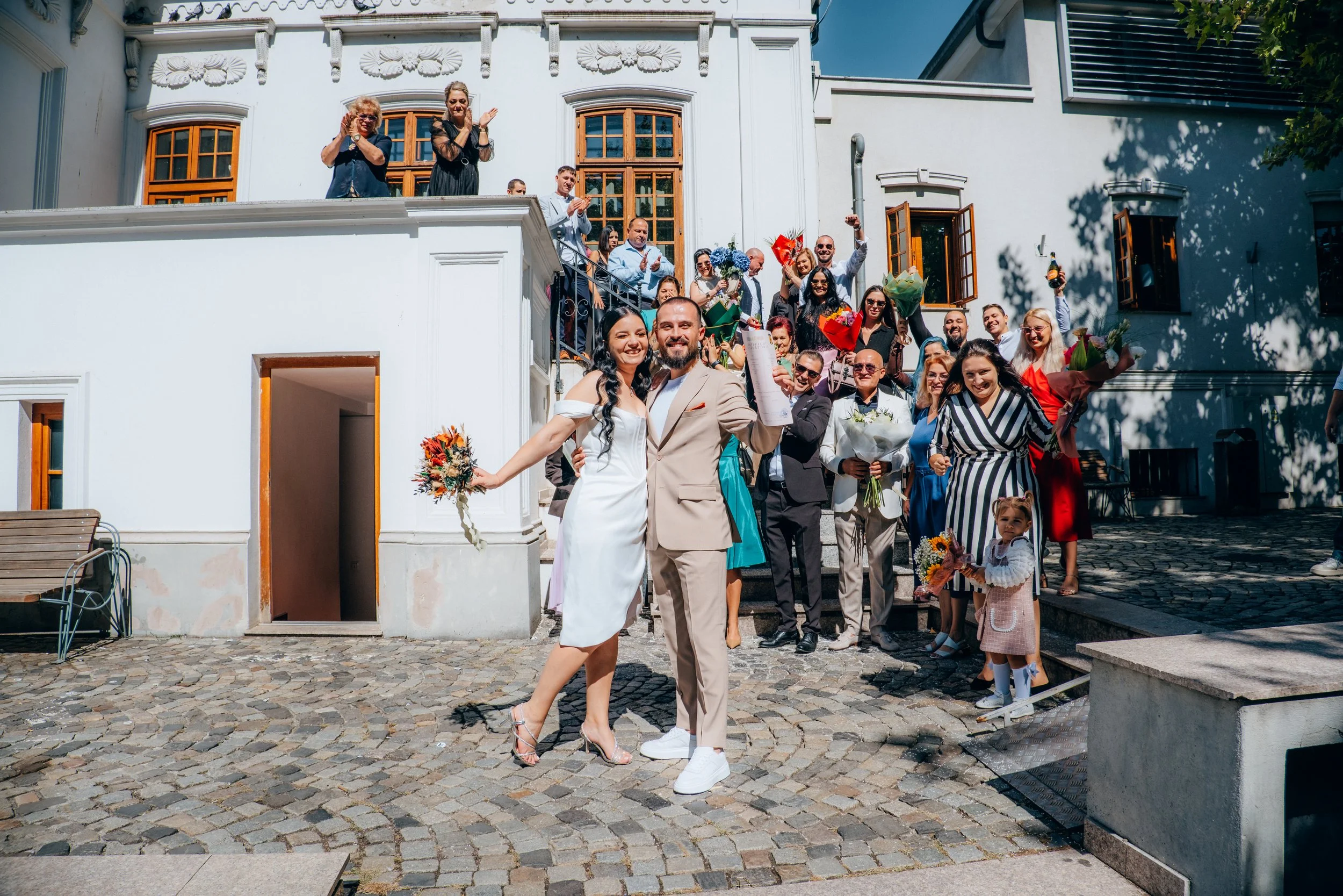 A wedding celebration outside a white building with large windows. The bride and groom are in the foreground, holding flowers and smiling at the camera. Guests are gathered around, cheering, clapping, and holding bouquets, with some cheering on a sta