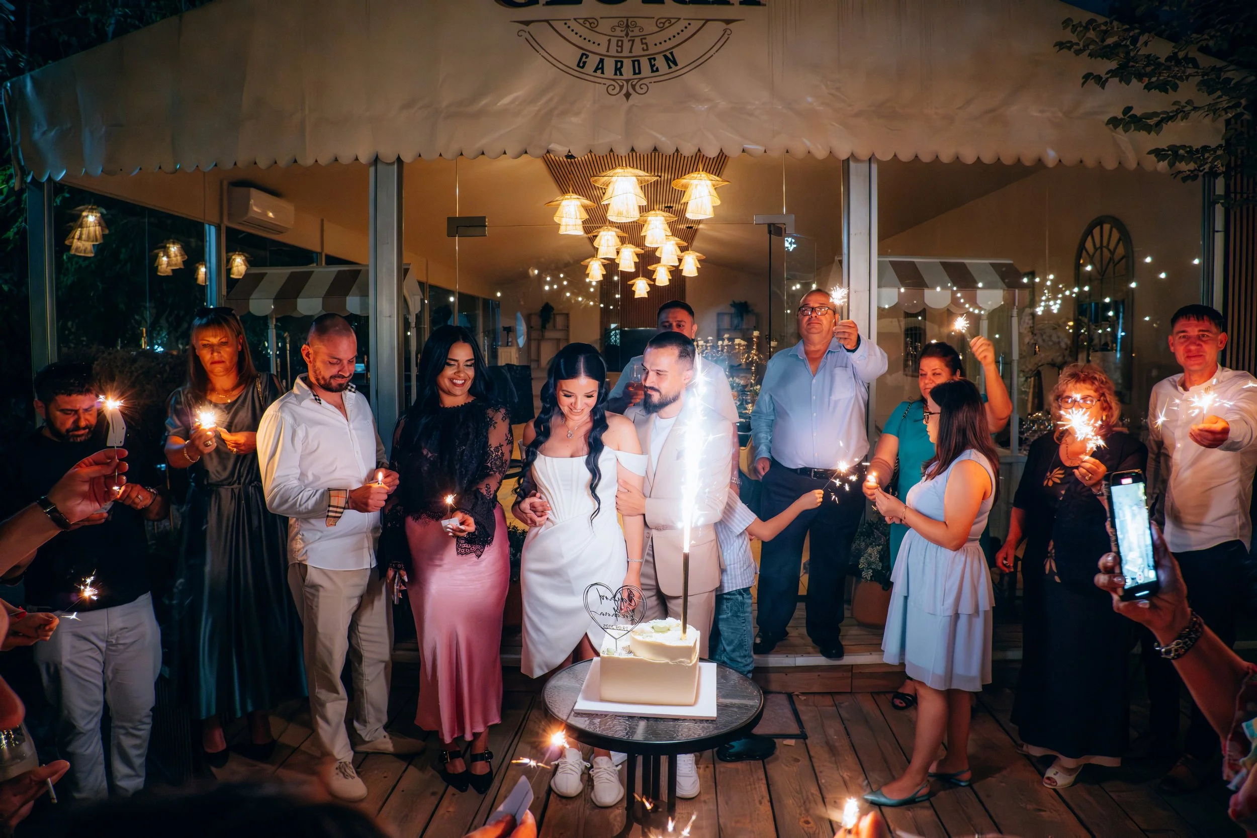 A group of people gathered under a canopy celebrating a birthday. They are holding sparklers and looking at a cake on a table. The scene is illuminated with warm lighting and decorated for a festive occasion.