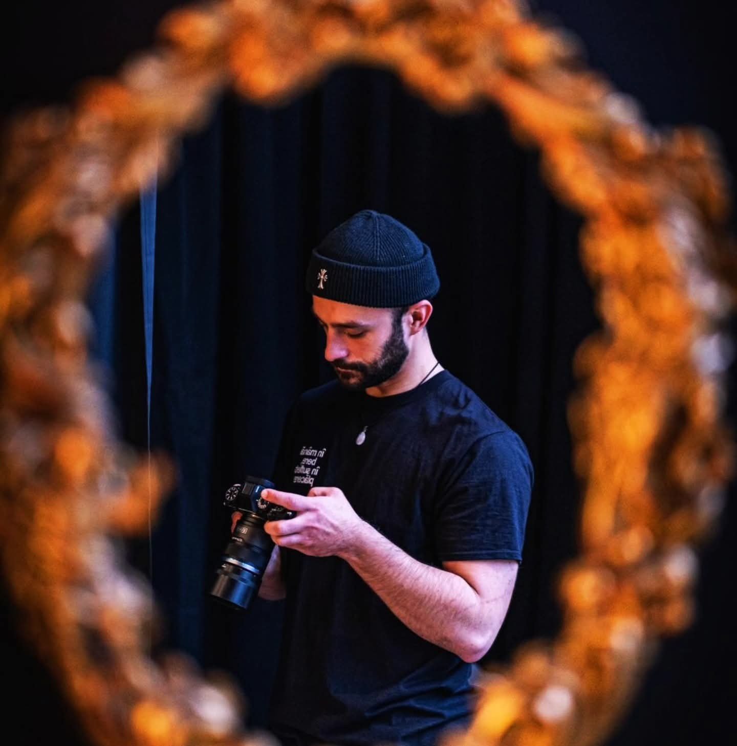 A man with a dark beard, wearing a black beanie and black T-shirt, looking at a camera he is holding in his hands, framed by an out-of-focus orange-brown decorative border.