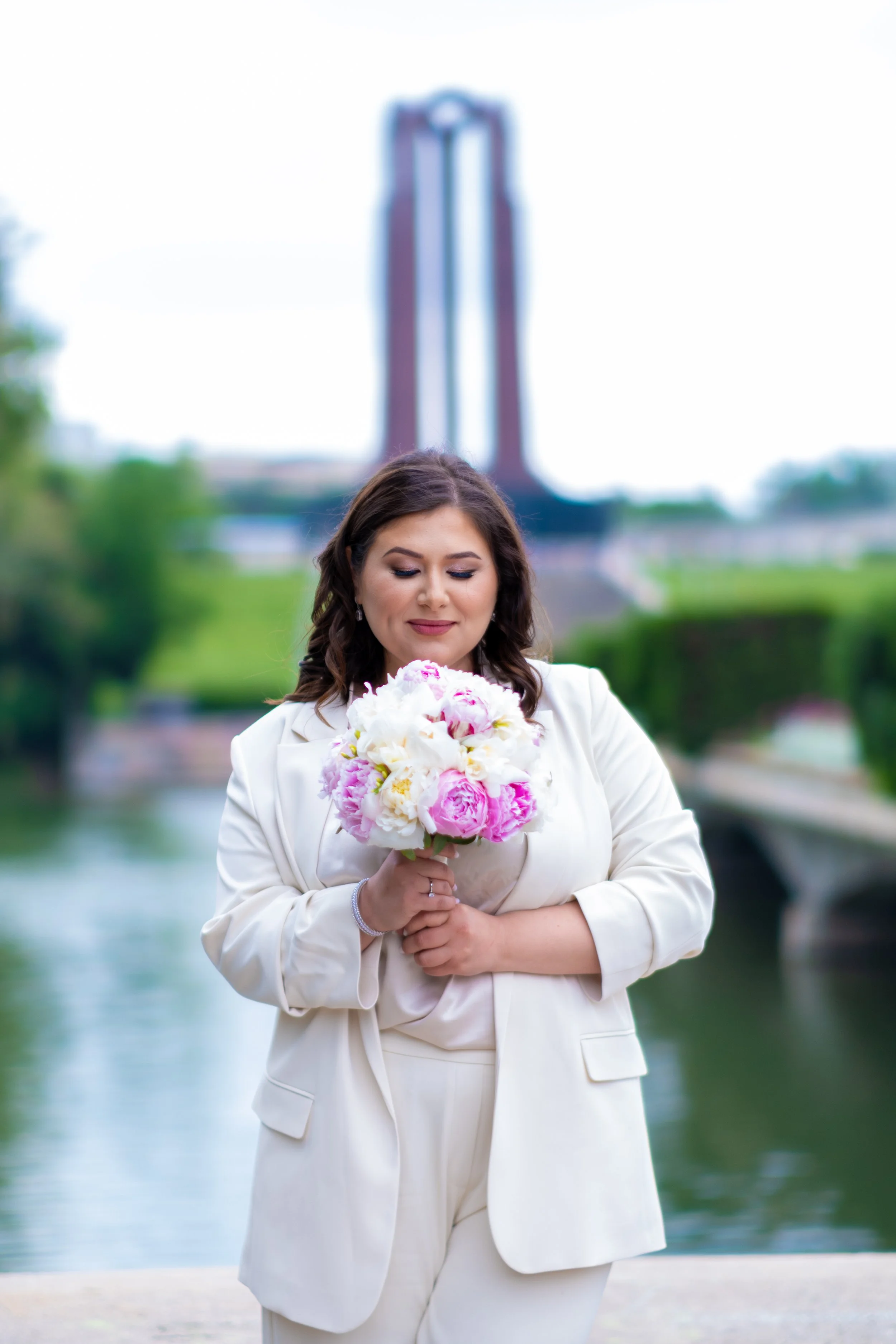 A woman in a white suit holding a bouquet of pink and white flowers, standing outdoors near a body of water with a bridge in the background.