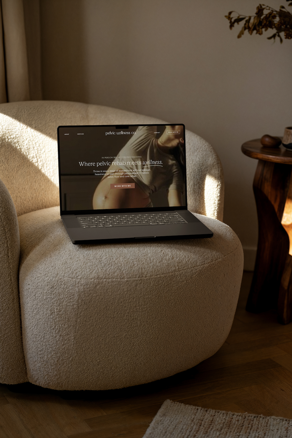 Laptop on beige armchair displaying a website about pelvic wellness and rehab, with sunlight casting shadows on the chair and a wooden side table nearby.