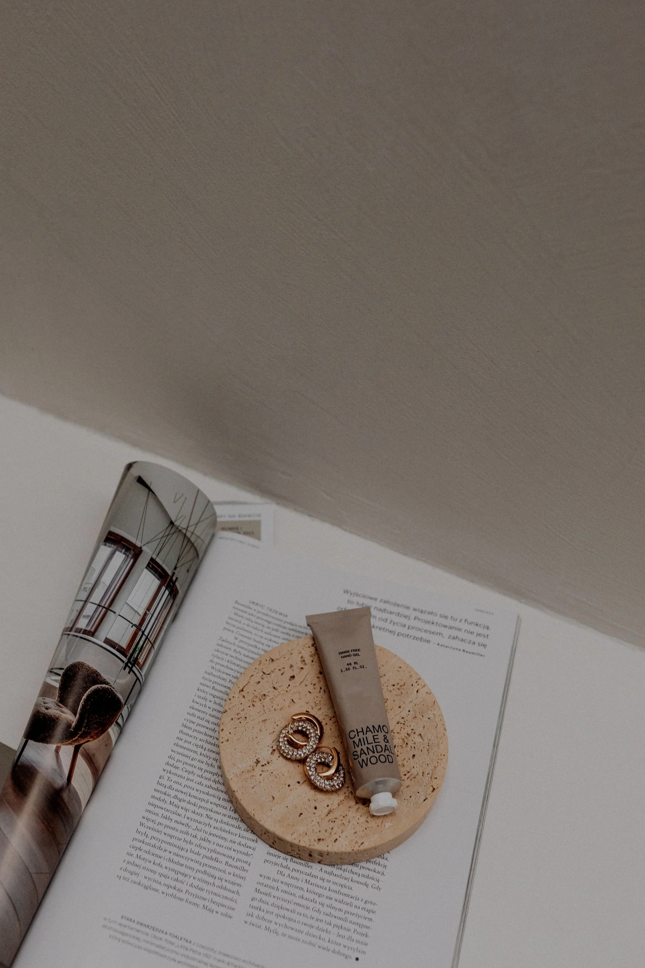 A beauty or skincare tube labeled 'Chamois & Sandalwood' resting on a round beige sponge, a pair of gold earrings with embedded stones, and an open magazine with wall-mounted lights and a chair on a white surface.