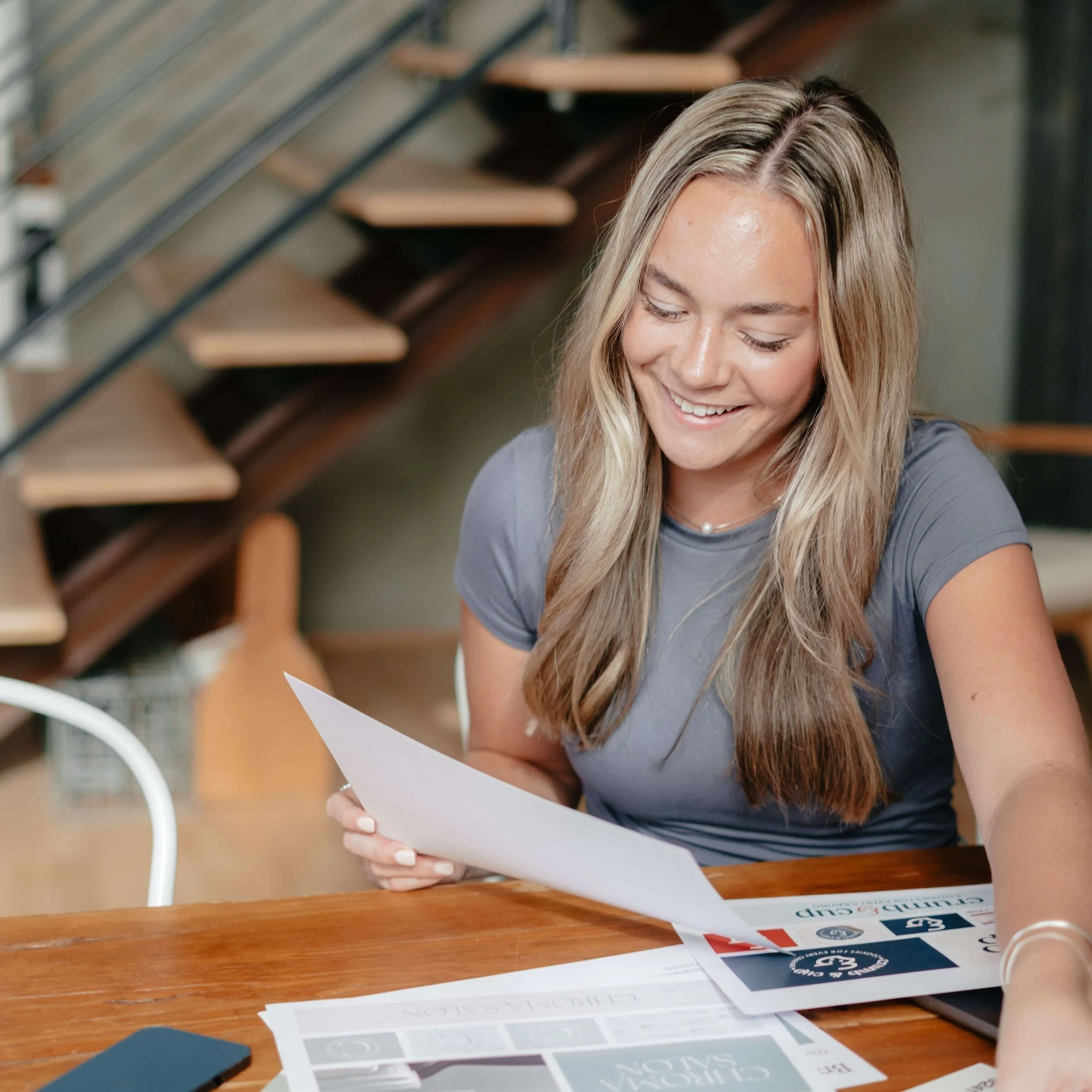 A young woman with long blonde hair, wearing a gray t-shirt and a pearl necklace, smiling while looking at a printed document at a wooden table inside a modern office or home workspace.