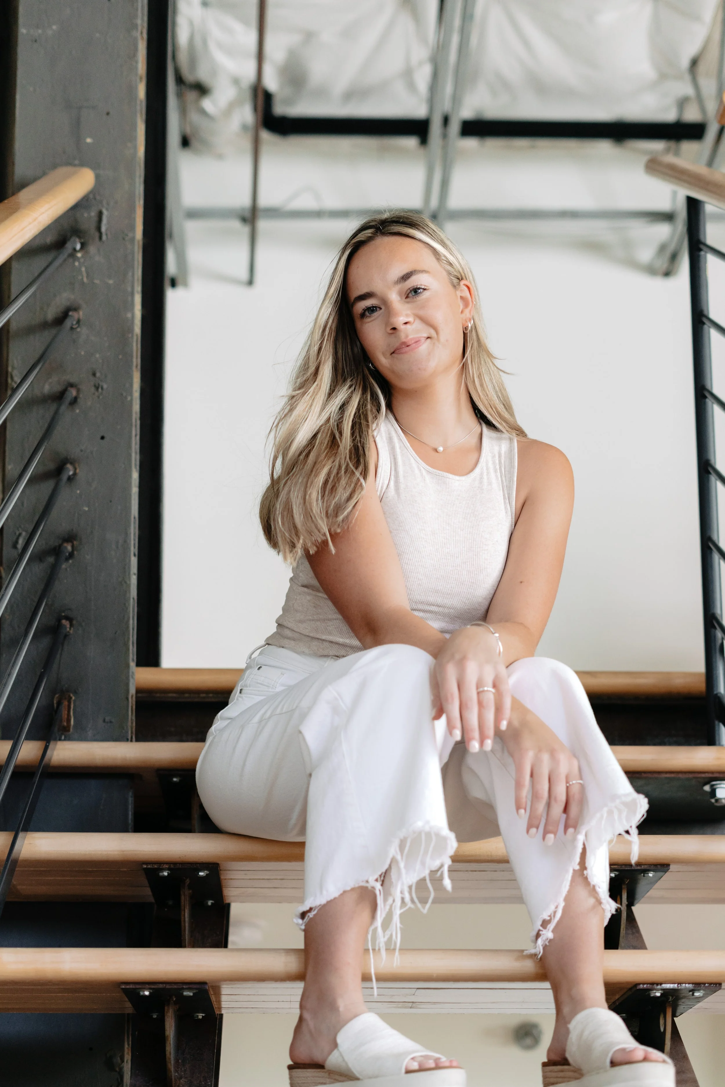 A young woman with long blonde hair sitting on a staircase with wooden steps in an industrial-style interior, wearing a beige sleeveless top and white frayed jeans, smiling softly at the camera.