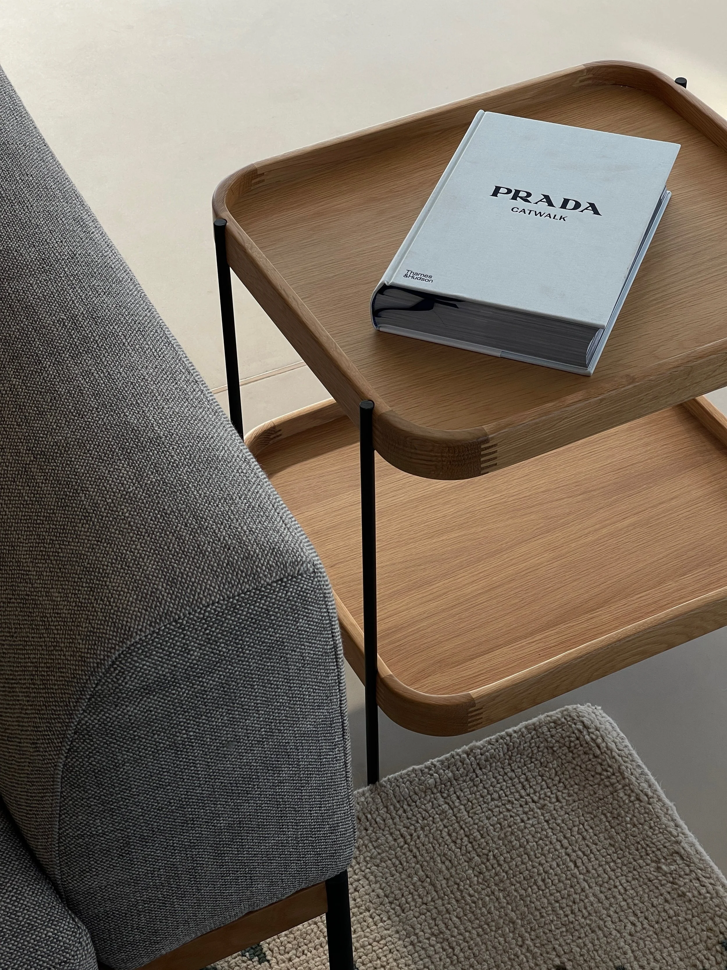 A wooden tray table with a Prada catalog resting on top, next to a gray upholstered chair, with a beige textured rug on the floor.