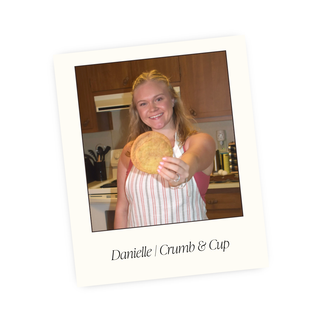A woman smiling and holding a large cookie in a kitchen setting.