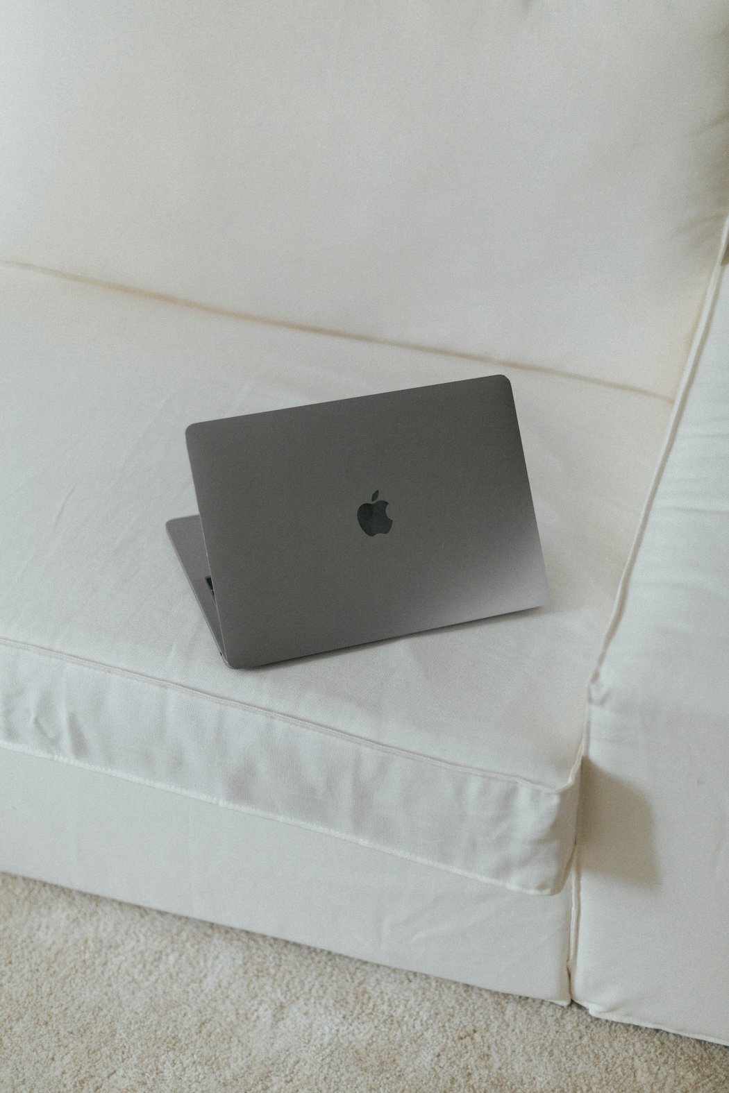 A closed silver MacBook laptop resting on a white bed with a white pillow in the background.