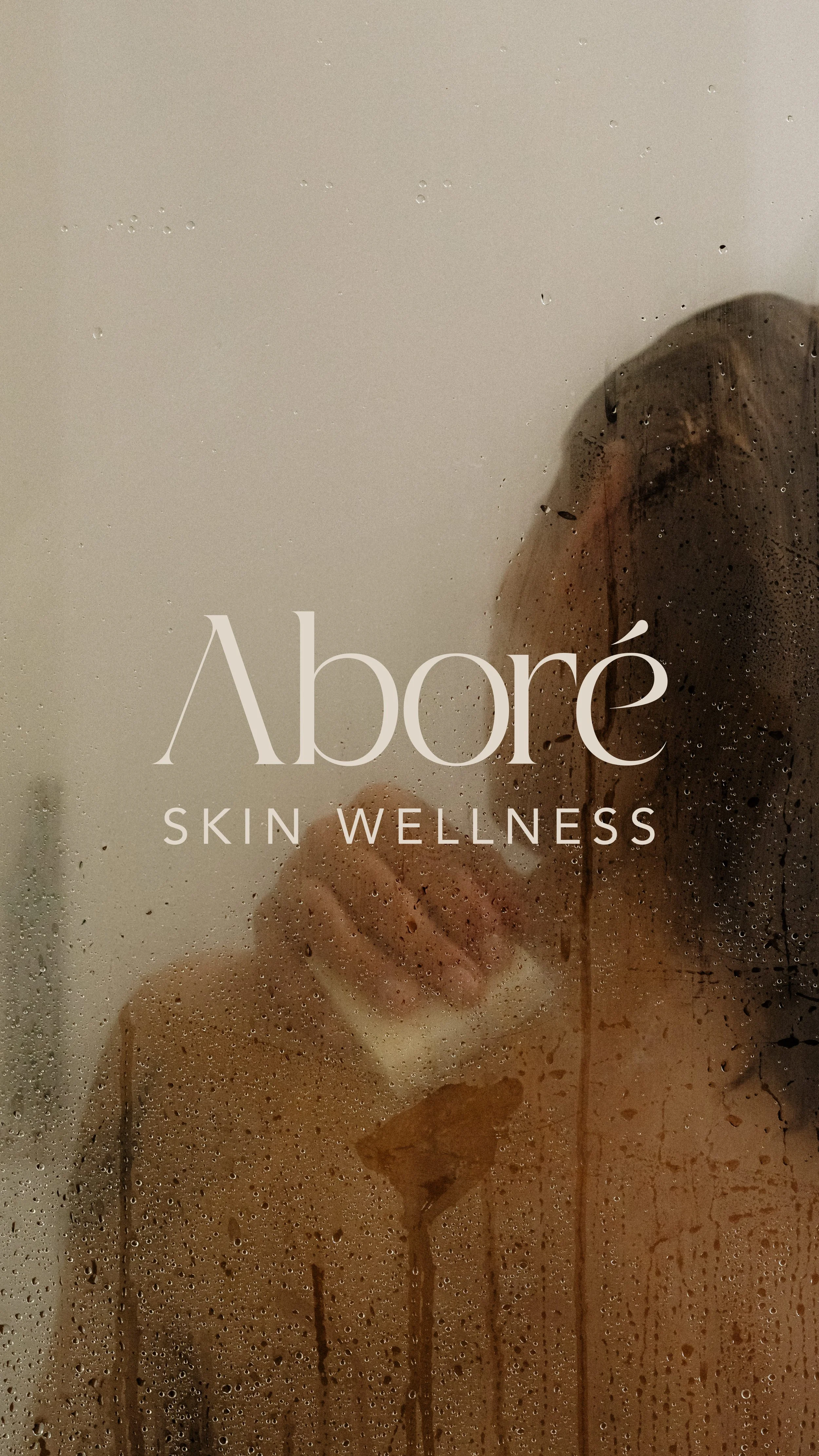A person with wet hair taking a shower behind a foggy glass door with water droplets, with text overlay reading 'Àboré SKIN WELLNESS'... the white logo for a passion project designed by Evelle Creative.