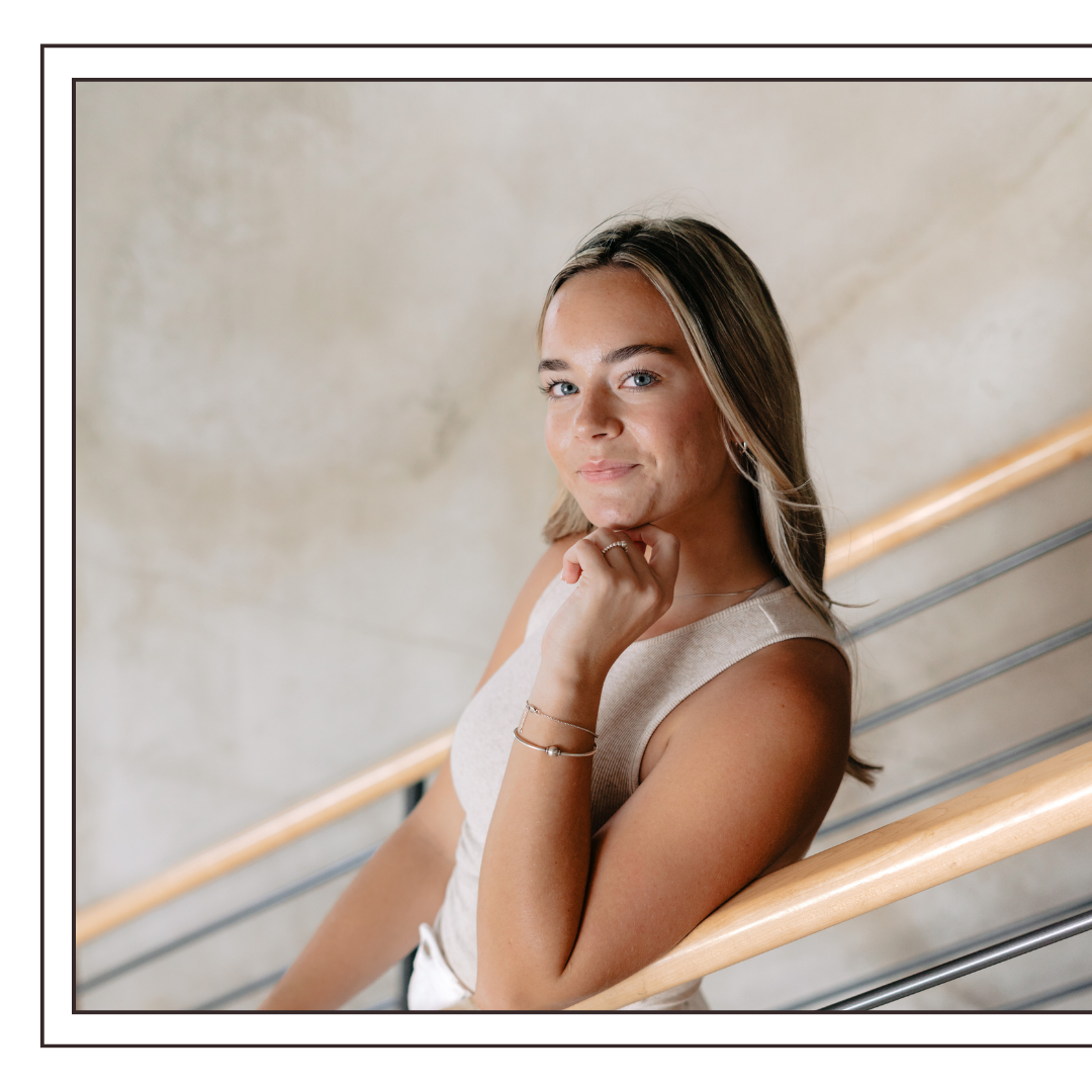 Abbey Delaney, founder of Evelle Creative, is wearing a sleeveless beige top, sitting on a staircase with wooden handrails, looking at the camera with a slight smile.