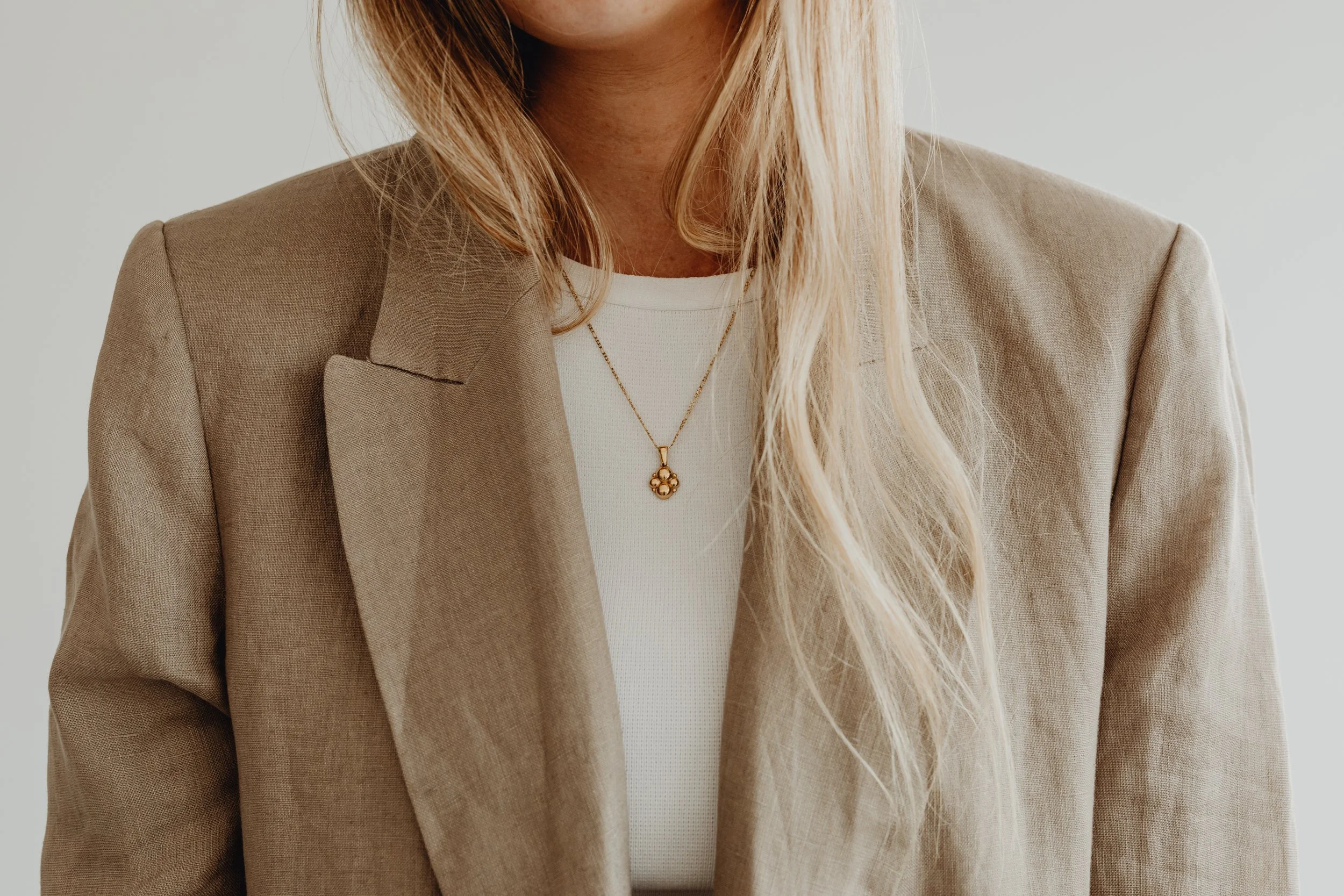 Close-up of a woman’s upper body, wearing a beige blazer, white top, and a gold necklace with a floral pendant.