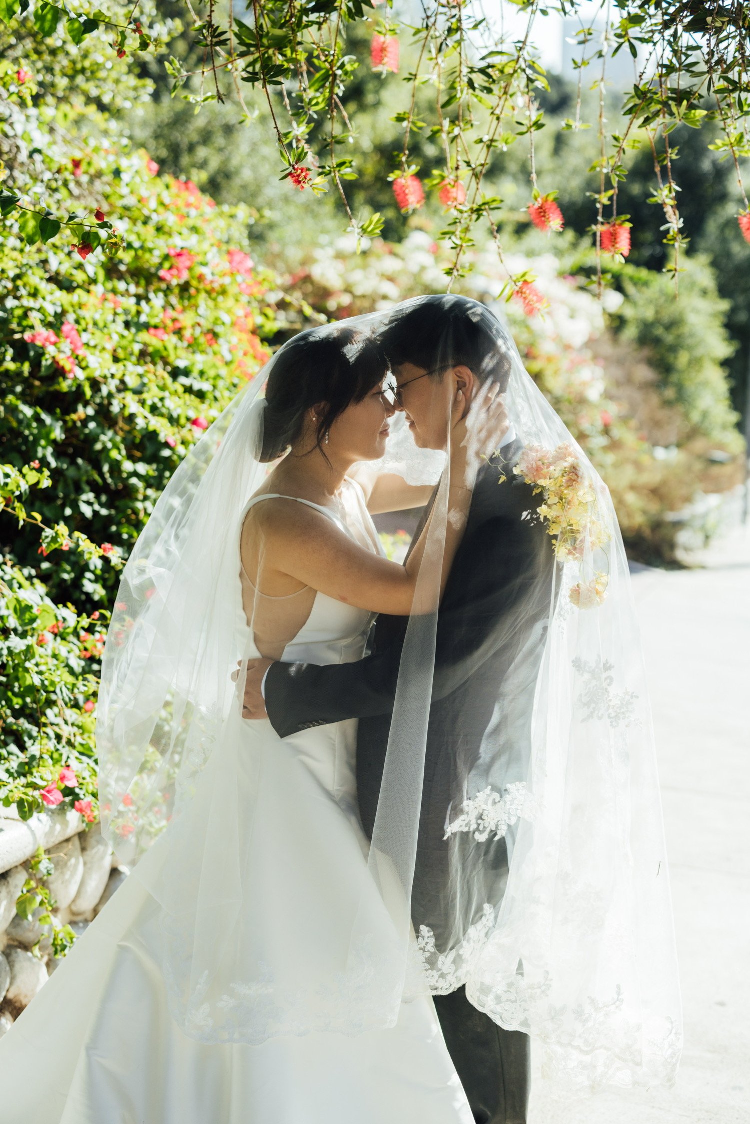 A bride and groom in wedding attire embrace under a veil, standing outdoors surrounded by lush greenery and blooming flowers.