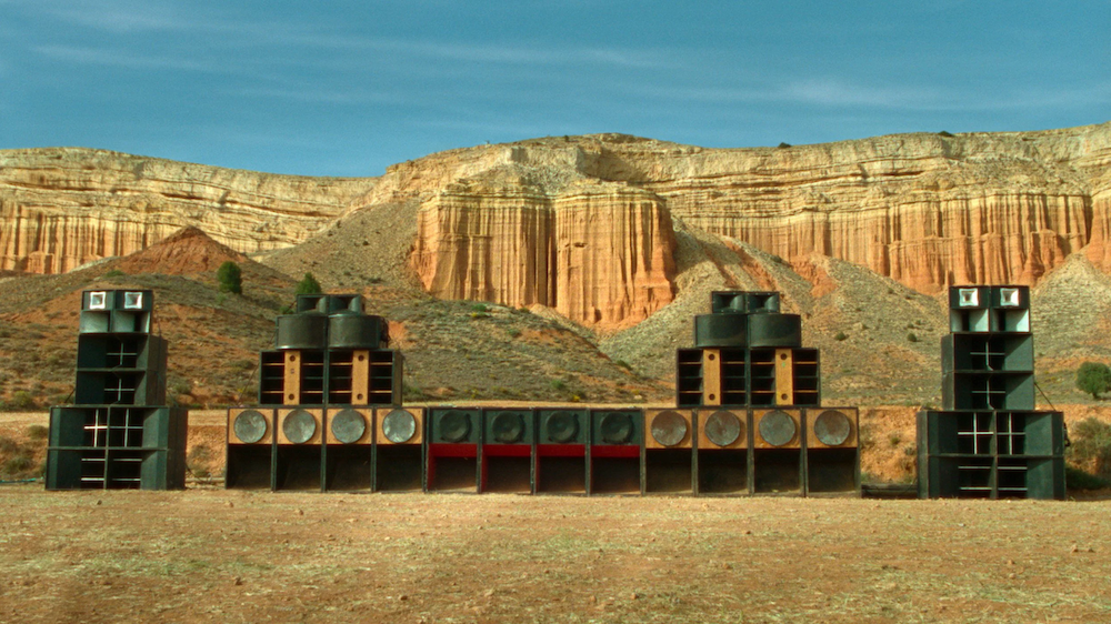 A film still from Sirat showing massive speakers and amps in a desert