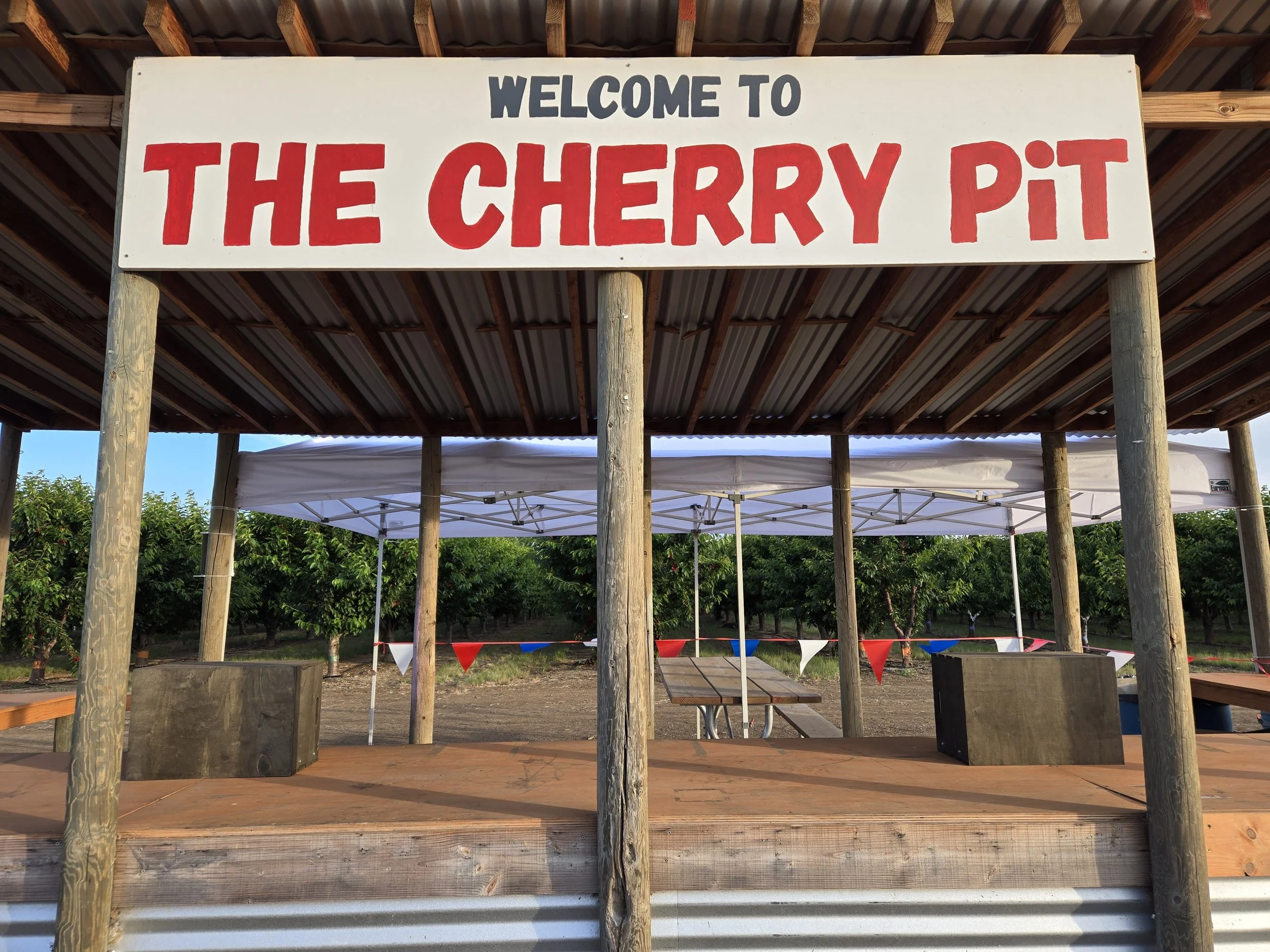 A sign hanging above a farm stand that reads 'Welcome to The Cherry Pit.' The stand is outdoors, with cherry trees and a tented canopy in the background.