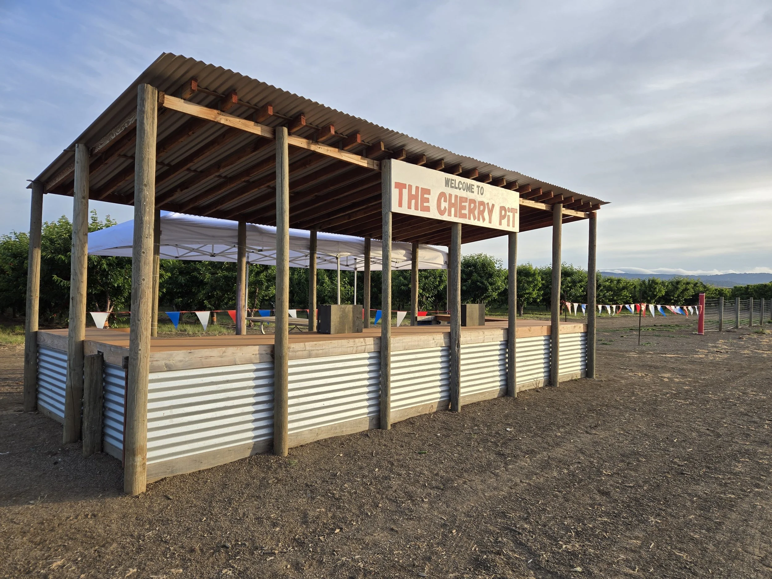 A farm stand with a sign that reads 'Welcome to The Cherry Pit.' The stand has a corrugated metal skirt, wooden support beams, and a metal roof. In the background, there are cherry trees, flags, and a distant fence and mountains.