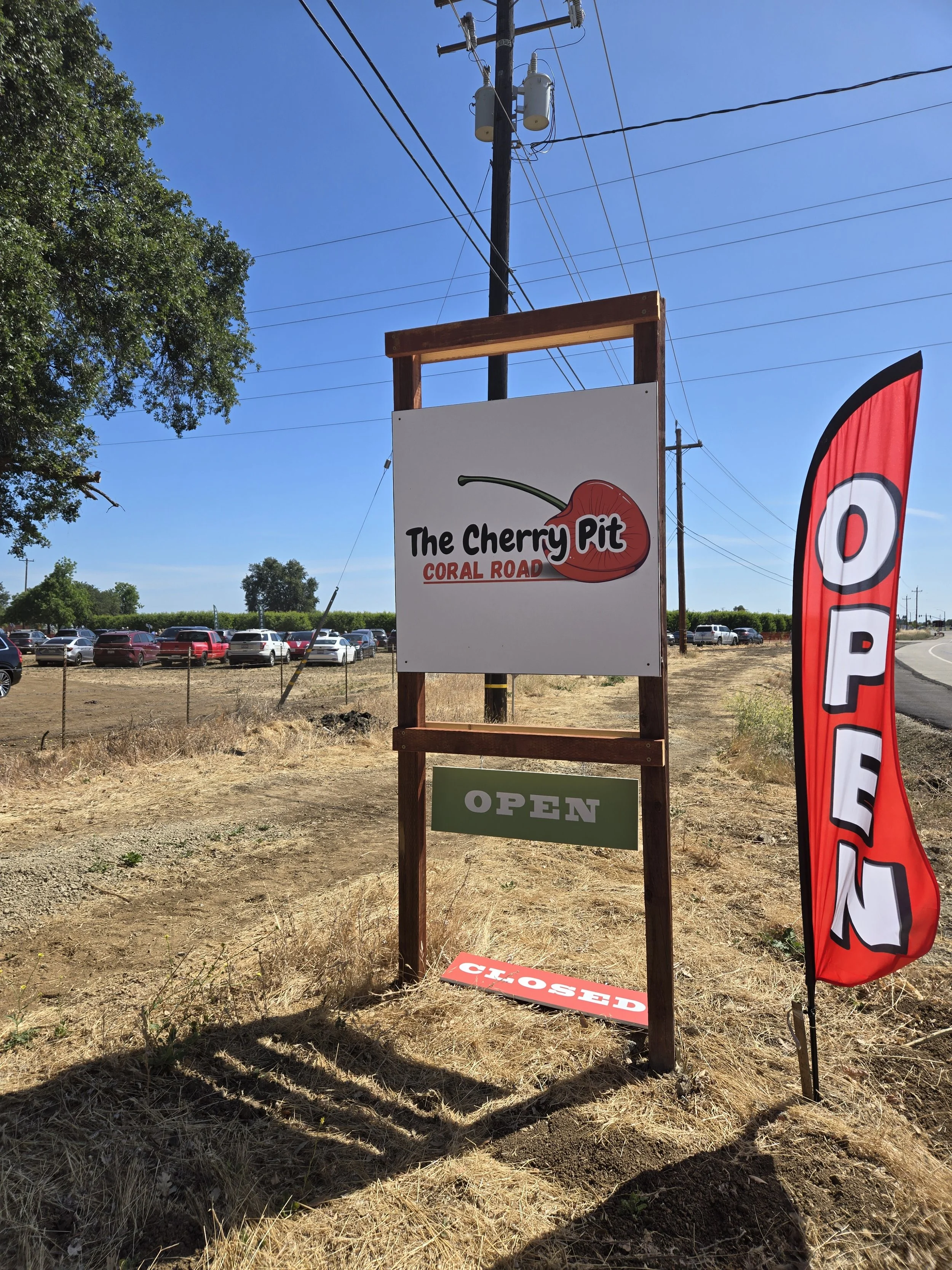 Sign for The Cherry Pit: Coral Road, the Cherry Pit's second location. There is a red and white flag with the word "OPEN" beside it. A parking lot with multiple cars is visible in the background under a clear blue sky.