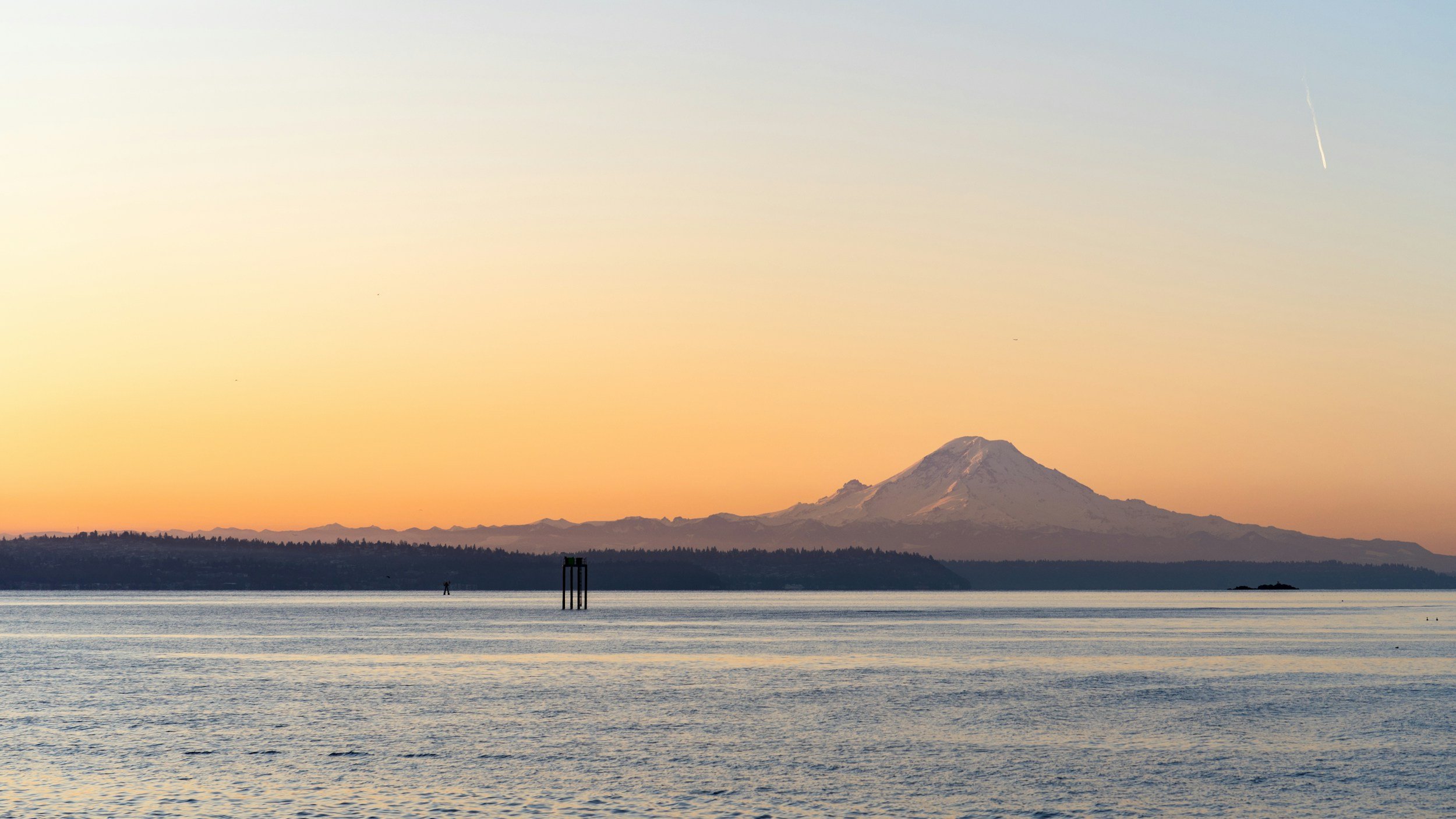 A view of a mountain, likely Mount Rainier, with snow cap, in the distance over a body of water during sunset, with a clear sky, some distant land, and a jet trail in the upper right corner.