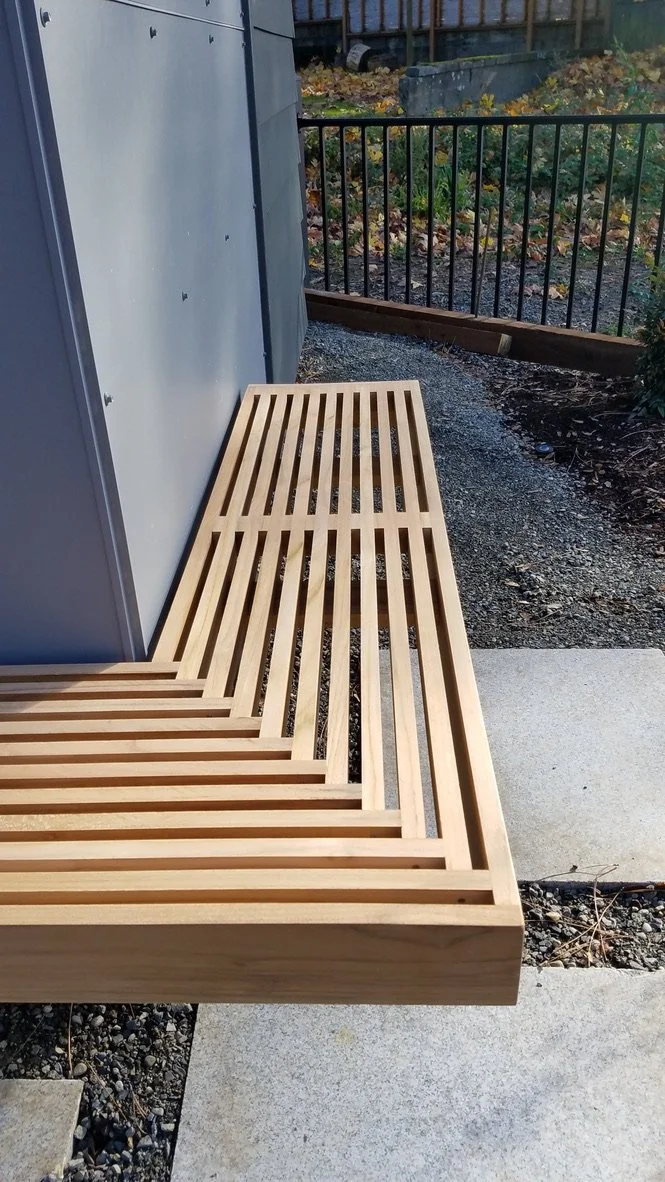 A wooden slatted bench with a backrest placed outdoors on a concrete and gravel surface next to a gray metal structure with a black metal fence and a garden in the background.