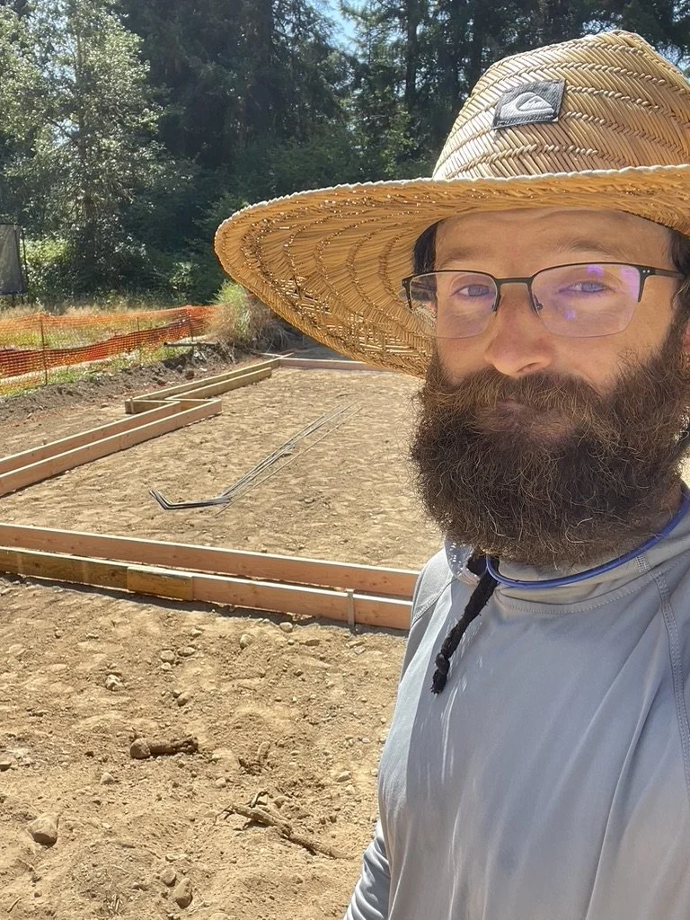 Man with glasses and a full beard wearing a wide-brimmed straw hat and a light gray long-sleeve shirt taking a selfie at a construction site with wooden framing for a sidewalk or pathway and trees in the background.